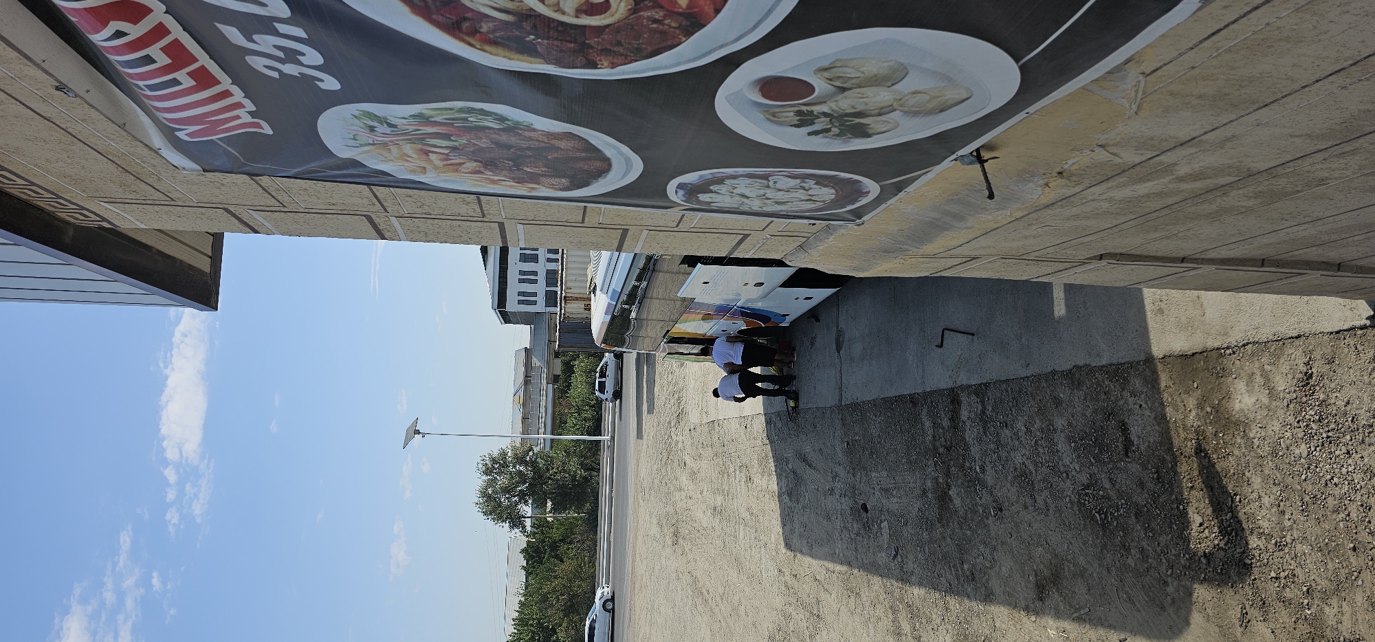 This is a photograph of a street scene in the city of Sheyhancha, Uzbekistan, on a clear, sunny day. In the foreground, a large banner advertising food is attached to a textured, light-colored building. The banner features images of various dishes, including what appears to be meat and dumplings. Two men, dressed in white shirts and dark pants, are working near the open doors of a bus parked on a paved area adjacent to the building. One man is bent over, appearing to be attending to the bus's wheels or undercarriage, while the other stands nearby. The bus has colorful decals on its side. A gravelly dirt area is to the left of the paved section, with tire tracks visible. Further in the background, across a road, is a tree-lined area with several buildings, including a modern white structure. A tall pole with a solar panel is also visible in the middle ground. Several cars are present on the road. The overall impression is one of a commercial or roadside service area.