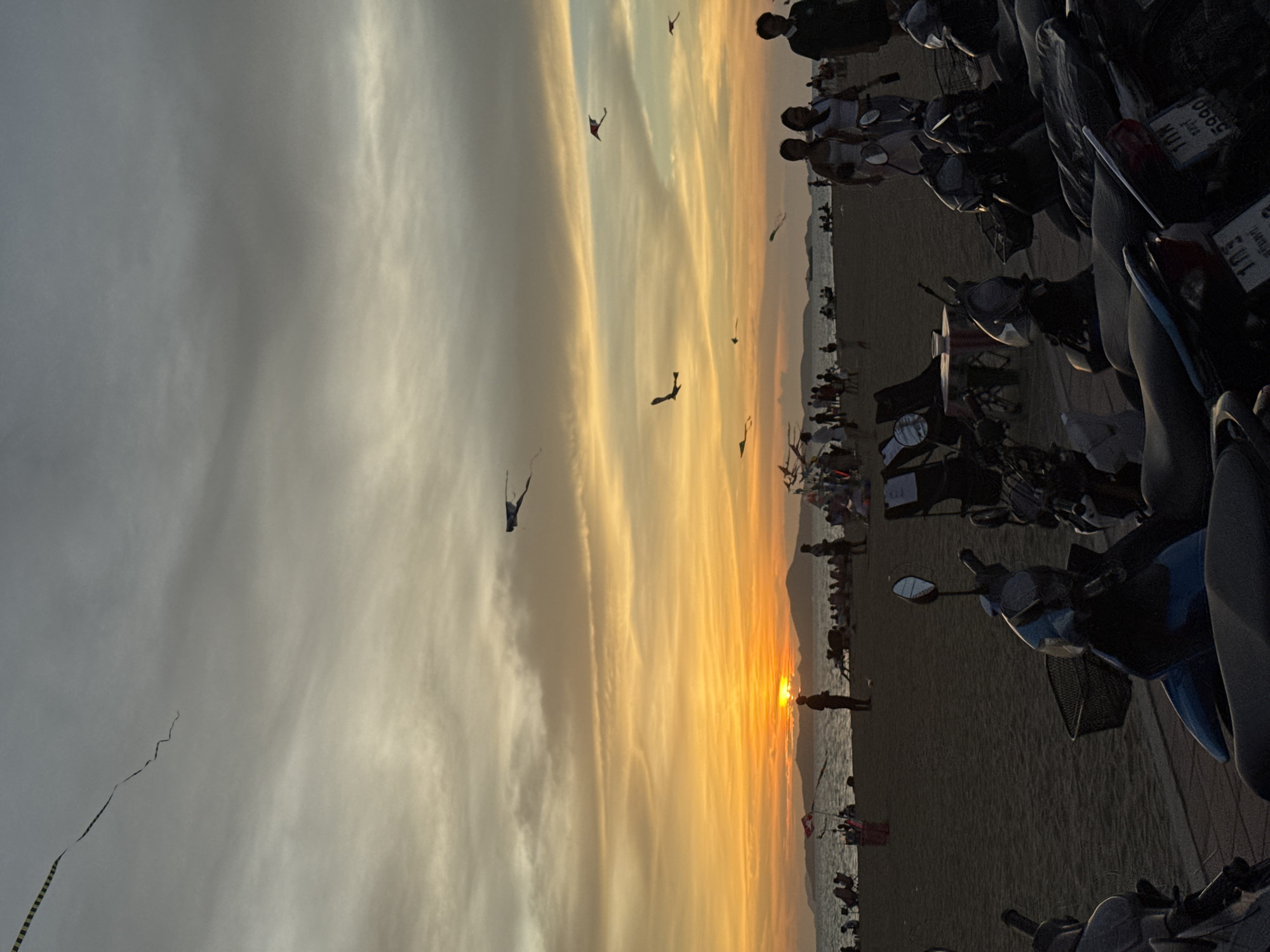 Captured in Pattaya City, Thailand, the image depicts a sunset scene on a beach, with a multitude of people flying kites. Motorbikes are parked on the periphery, and the overall mood is serene.