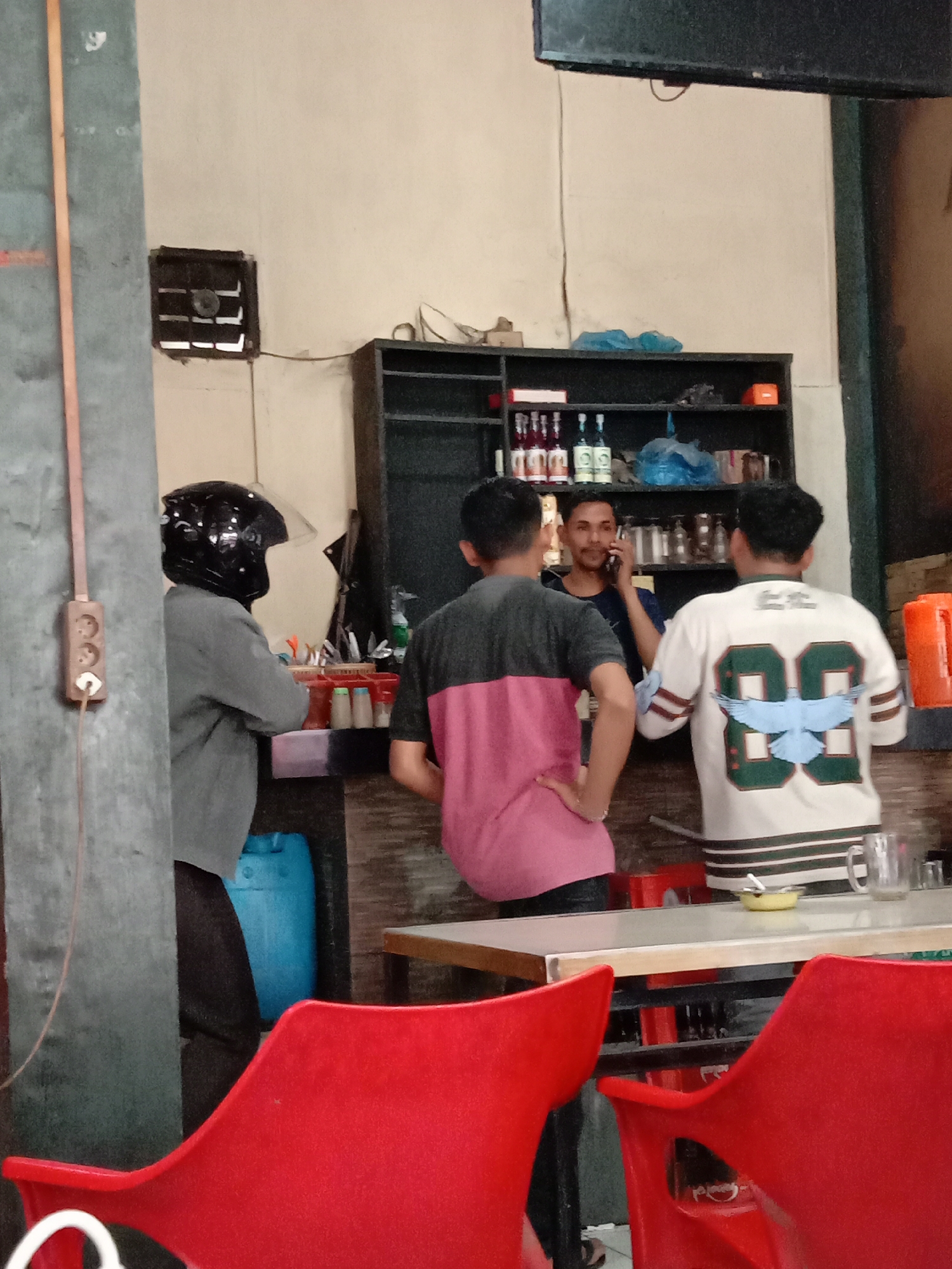 The image captures an indoor scene, likely a roadside eatery or warung, in Lhokseumawe, Indonesia. Three men are present. On the left, a person wearing a black motorcycle helmet and a gray long-sleeved shirt stands with their back to the camera, facing a counter. To their right, a man in a black t-shirt is on the phone, standing behind the counter. To the right of the man on the phone, another man is seen from the back, wearing a white t-shirt with green accents and the number "88" with a blue bird graphic on the back.
The environment includes a shelf unit behind the counter stocked with various bottles and containers, a blue plastic drum to the left of the counter, and a table with red chairs in the foreground. The walls appear to be a neutral color, possibly cream or off-white, and a light fixture or speaker is mounted on the ceiling. The overall impression is of a casual, functional establishment.
