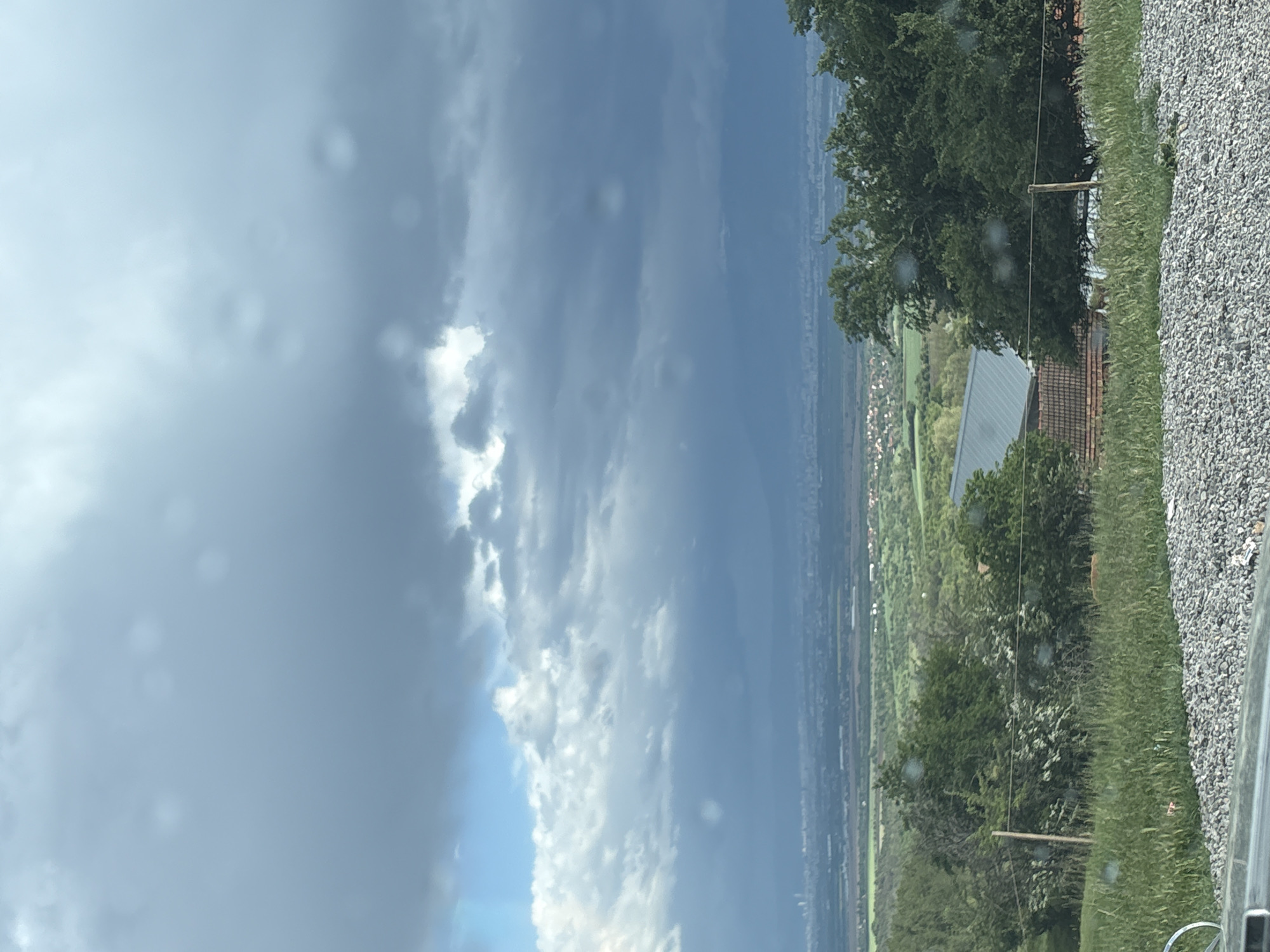 The image shows a view from an elevated position, possibly from a moving vehicle, overlooking a landscape with a dramatic sky. The sky is dominated by dark, heavy clouds with patches of bright white clouds and blue sky breaking through. The foreground appears to be part of a vehicle window with water droplets on it.
Below the sky, there is a panoramic view of a valley or plain, featuring a mix of greenery, fields, and what seems to be a settlement or urban area in the distance. In the mid-ground, there are trees and what looks like a building with a gray roof and reddish-brown tiles visible. The immediate foreground consists of roadside vegetation and a gravelly surface.
There are no people visible in the image. The overall environment suggests a rural or semi-rural setting with a visible storm or inclement weather in the area.