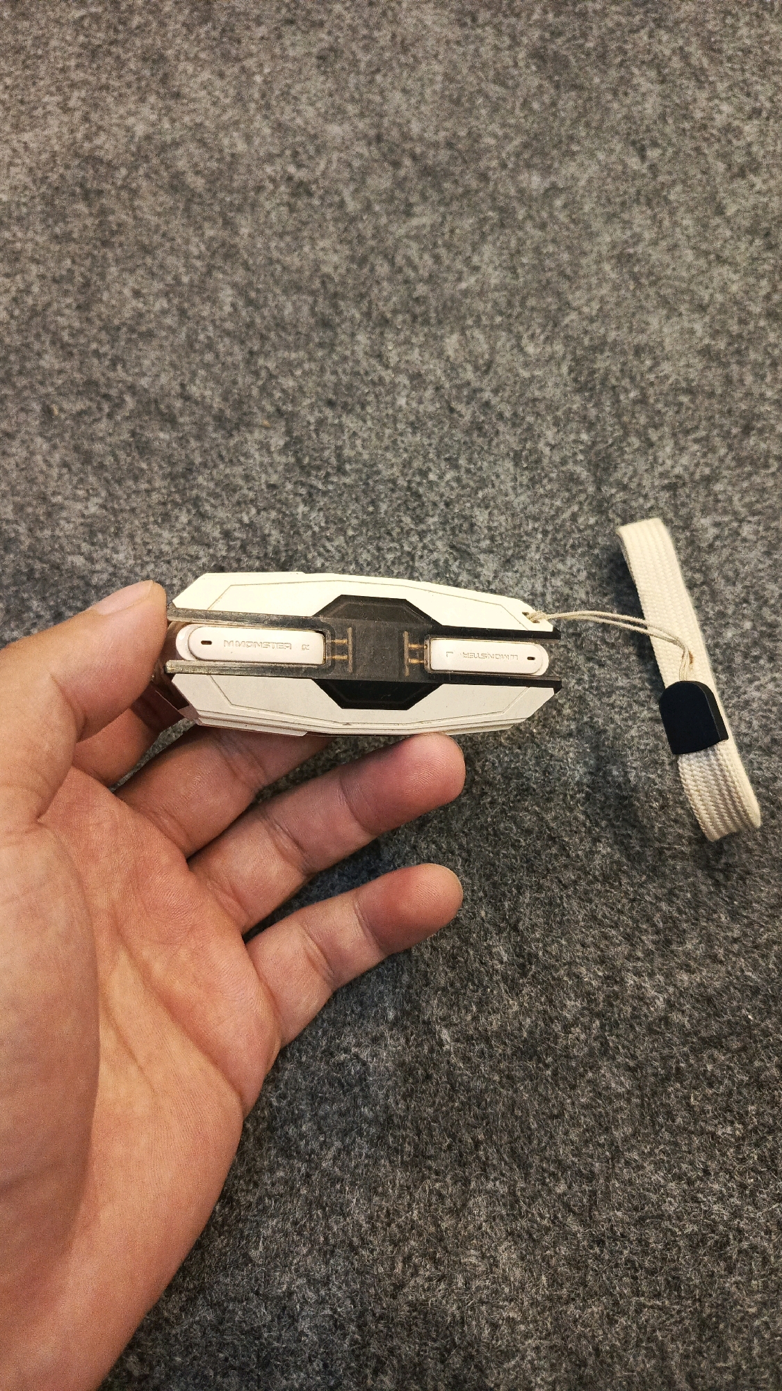 A left hand holds a white and black electronic device with a white wrist strap attached. The device has text that reads "MMONSTER R" on the left side and "MMONSTER L" on the right side. The device is resting on a gray, textured surface, likely a carpet or felt material. There are no visible elements to indicate the location in the city of Babadan, Indonesia.