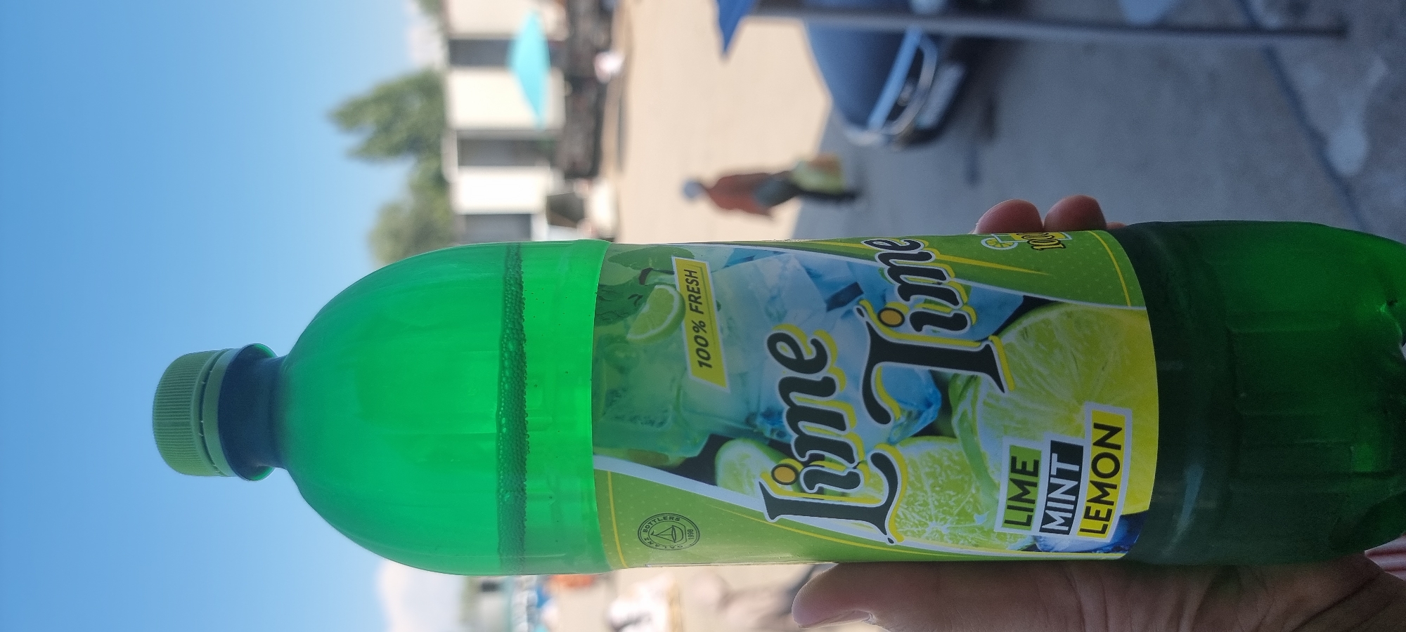 A person's hand holds a green plastic bottle of "Lime Time" flavored drink, which features an image of limes and ice on its label. The bottle has a green cap and appears partially filled with a dark green liquid, with some bubbles visible near the top. The label also indicates "100% FRESH" and lists "LIME MINT LEMON" as flavors. The background suggests an outdoor setting, possibly a public area or marketplace, with blurred figures of people and buildings. The overall scene is brightly lit by daylight. The sky is clear and blue.