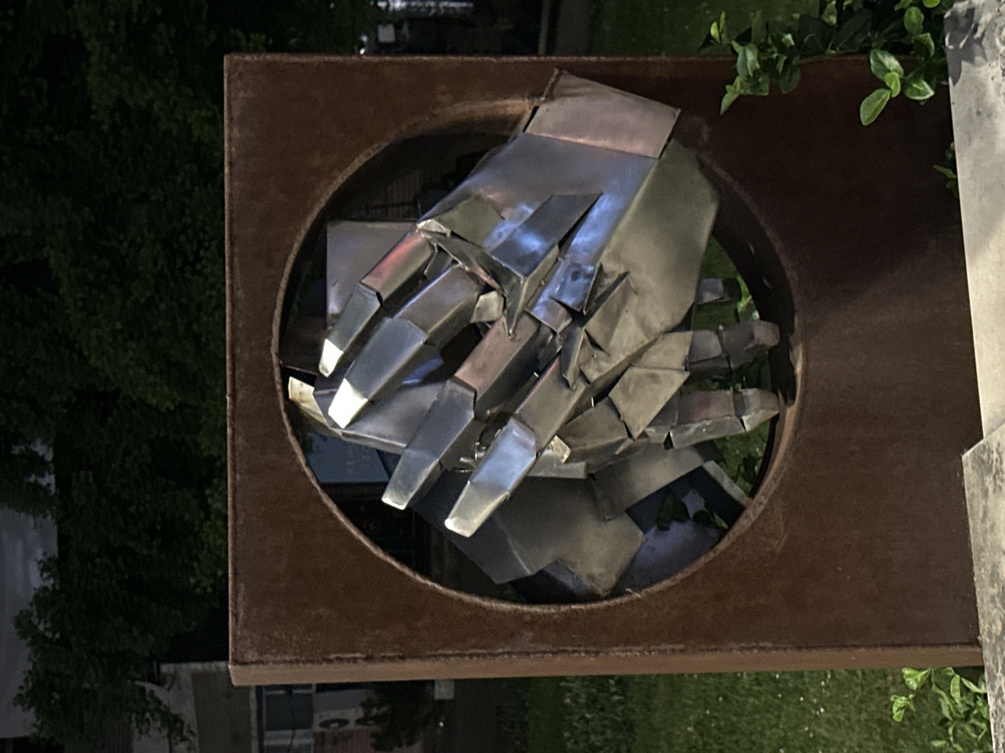 An outdoor sculpture in Veliko Tarnovo, Bulgaria. The sculpture is of two hands made of silver colored metal within a circular cutout of a rusted metal frame, set in a square rusted metal frame. The metal frame is positioned on what appears to be a stone pillar. The surroundings show nighttime foliage with dark green leaves and a building in the distance.