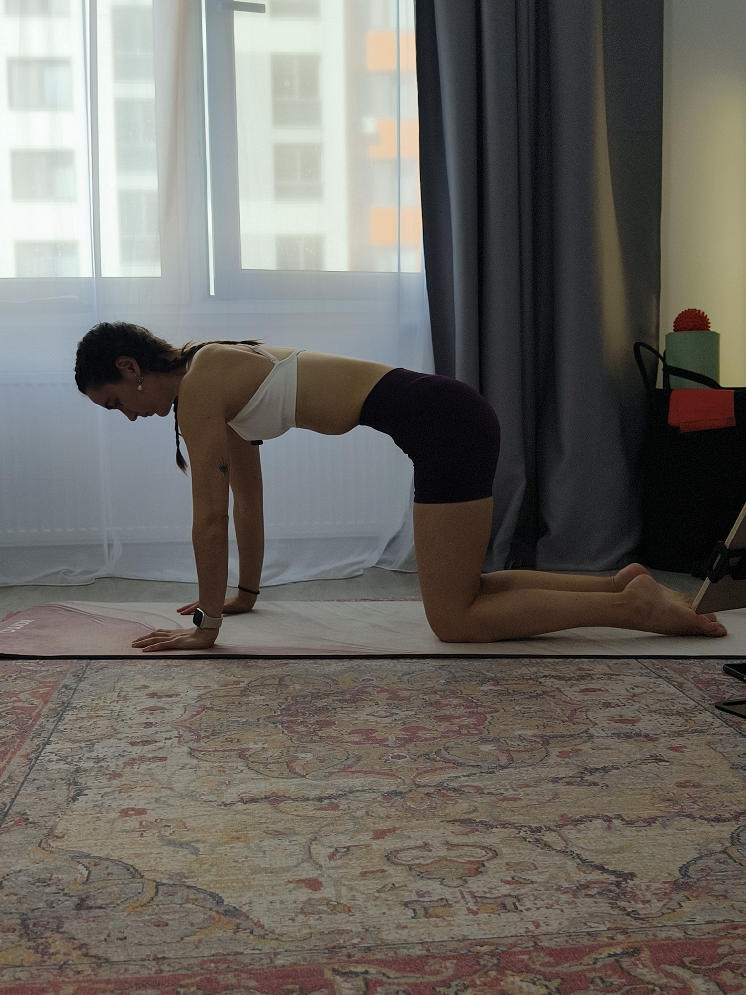 A woman is in a tabletop position on a yoga mat in a room. She is facing left and is on her hands and knees. Her back is arched upwards and her abdomen is pulled inwards. She is wearing a white sports bra and dark purple shorts. Her hair is pulled back in a braid. A patterned rug is on the floor in front of the mat. Behind her is a window with sheer curtains and a building visible outside. To the right, there is a dark gray curtain and a black bag with some fitness equipment. The scene appears to be indoors in a residential setting. The location is the city of Tyumen, Russia.