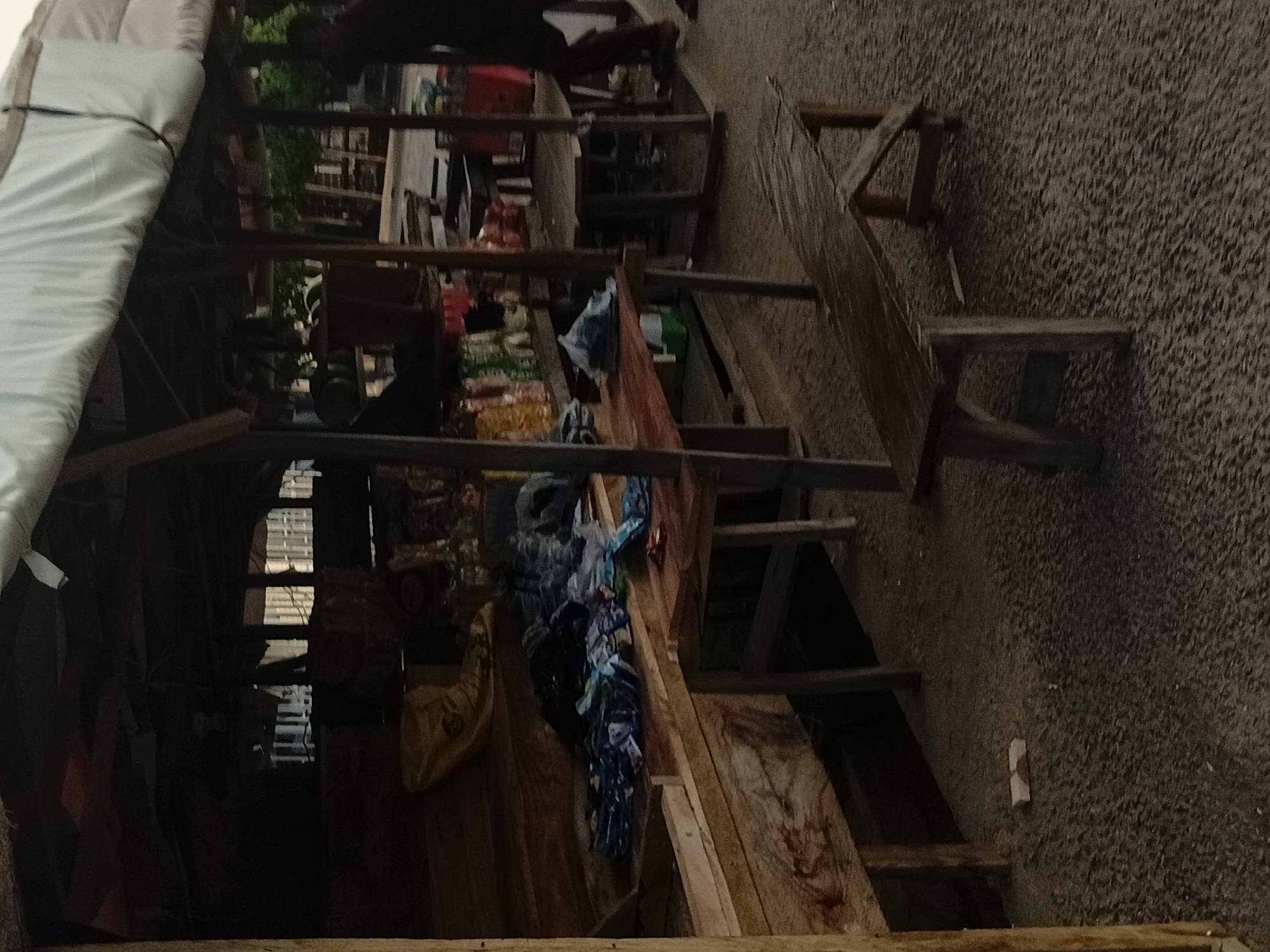 The image depicts an outdoor market stall in Jalingo, Nigeria. The stall is constructed with wooden beams and covered with a white tarp. Several wooden tables are set up, displaying various goods, including packaged snacks and what appears to be fabric. A wooden bench is positioned in front of the stall. The ground is covered with gravel. The scene appears to be dimly lit, suggesting it might be early morning or late evening. The image is somewhat blurry and lacks sharp detail.