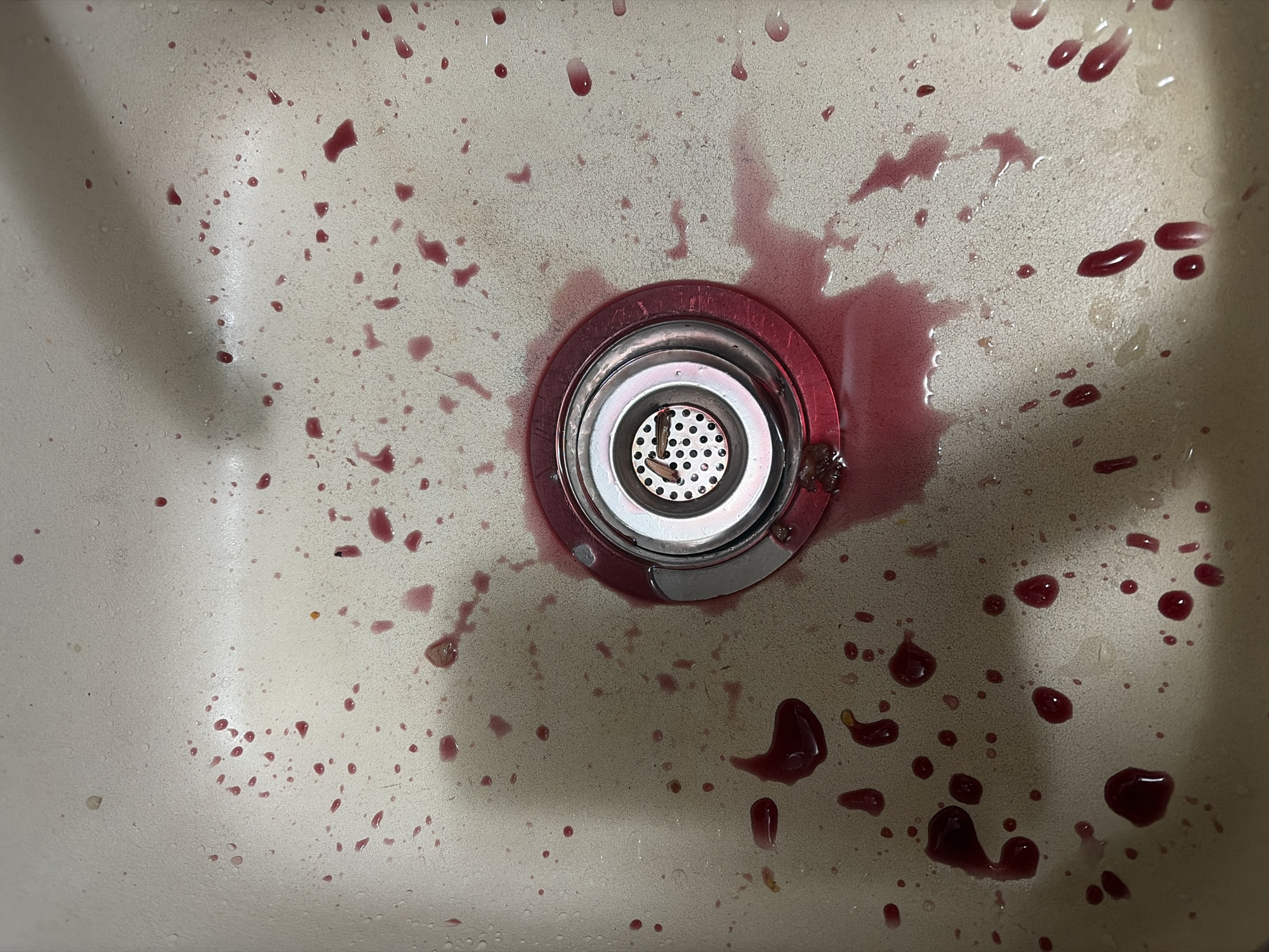 The image shows a close-up, overhead view of a sink basin, likely in Budva, Montenegro. The basin is a light, speckled color, possibly composite or ceramic. A chrome sink drain with a red rim is visible in the center, filled with a small amount of liquid. The basin is splattered with numerous droplets and streaks of a dark red liquid, consistent with fruit juice or wine. Some of these droplets are small and discrete, while others have coalesced into larger puddles. The overall impression is one of recent use and a minor spill.