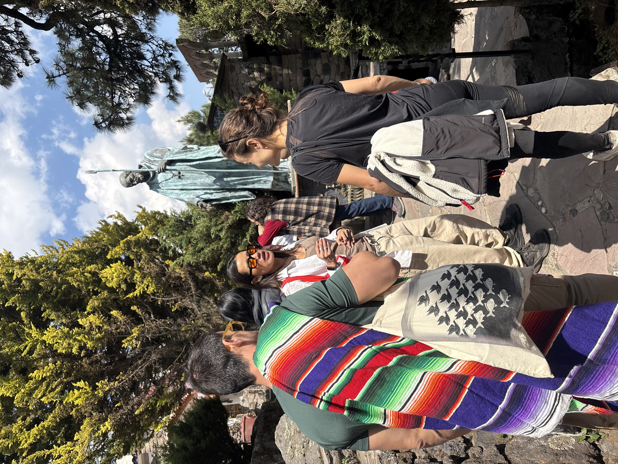 The image captures a gathering of several people in the city of San Mateo Ozolco, Mexico. They are situated near a stone statue depicting a robed figure holding a staff, possibly a religious or historical figure. The group appears to be resting or socializing on a stone pathway. One person is wearing a brightly colored, striped shawl, while another is draped with a light-colored cloth featuring a pattern of birds and fish. A woman stands wearing a black shirt and leggings with a jacket, looking towards the gathering. The backdrop includes trees and a partly cloudy sky. The environment suggests an outdoor setting, potentially a park or historical site.