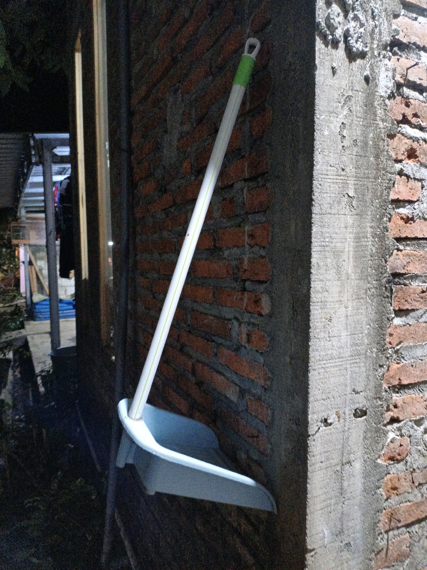 A dustpan and brush are leaning against a brick wall. The dustpan is light blue and the brush is white with a green handle. The brush is positioned above the dustpan. The wall is made of red bricks and has a rough, unfinished texture. There is a concrete pillar to the right of the dustpan and brush. The scene appears to be outdoors, possibly in a courtyard or alleyway. In the background, there are some stairs and a fence, suggesting a residential area. The lighting is dim, indicating it might be evening or nighttime. The image has a grainy quality. The location is Cikal, Indonesia.