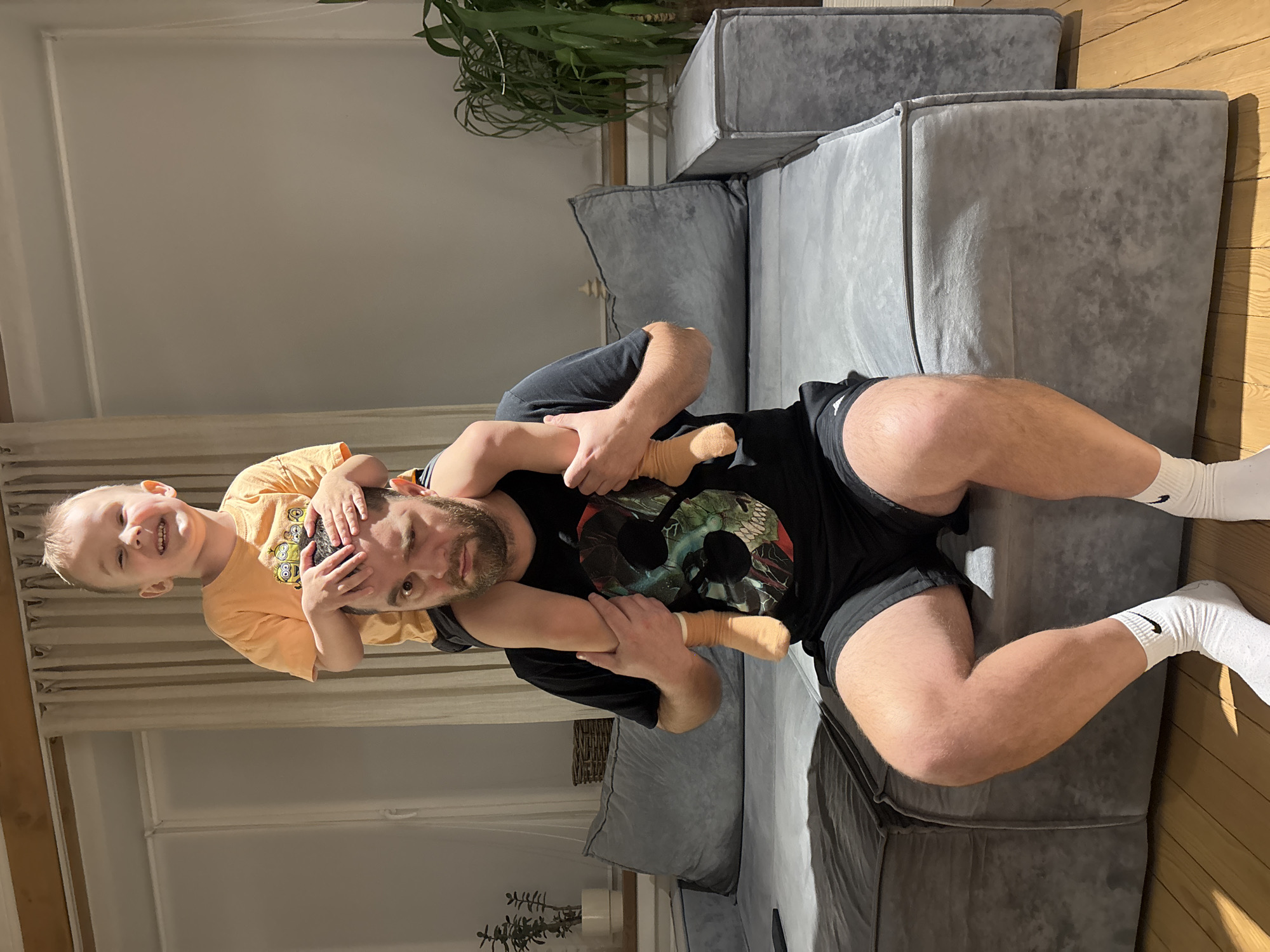 A heartwarming indoor photograph, taken in Chaikovsky, Russia, captures a playful moment between a man and a young boy. The man, likely the father, is positioned horizontally on his back on a light wooden floor, facing upwards with a content expression. He wears a black t-shirt featuring a complex graphic design, black shorts, and white socks with visible "Nike" branding.
Perched on the man's chest or stomach is a young boy, holding onto the man's head with both hands. The boy, with blond hair and a wide, joyful smile, is dressed in a bright orange t-shirt adorned with "Minions" characters and light-colored socks. His cheerful demeanor indicates a happy and engaged interaction with the man.
The scene takes place in what appears to be a living room, characterized by a large, modular gray fabric sofa occupying a significant portion of the background. Bright, natural sunlight streams in from an unseen window to the left, casting distinct shadows and illuminating the subjects and their surroundings. A green potted plant is visible in the upper background, and white baseboards line the walls. The image conveys a sense of domestic joy and affectionate family interaction during daylight hours.