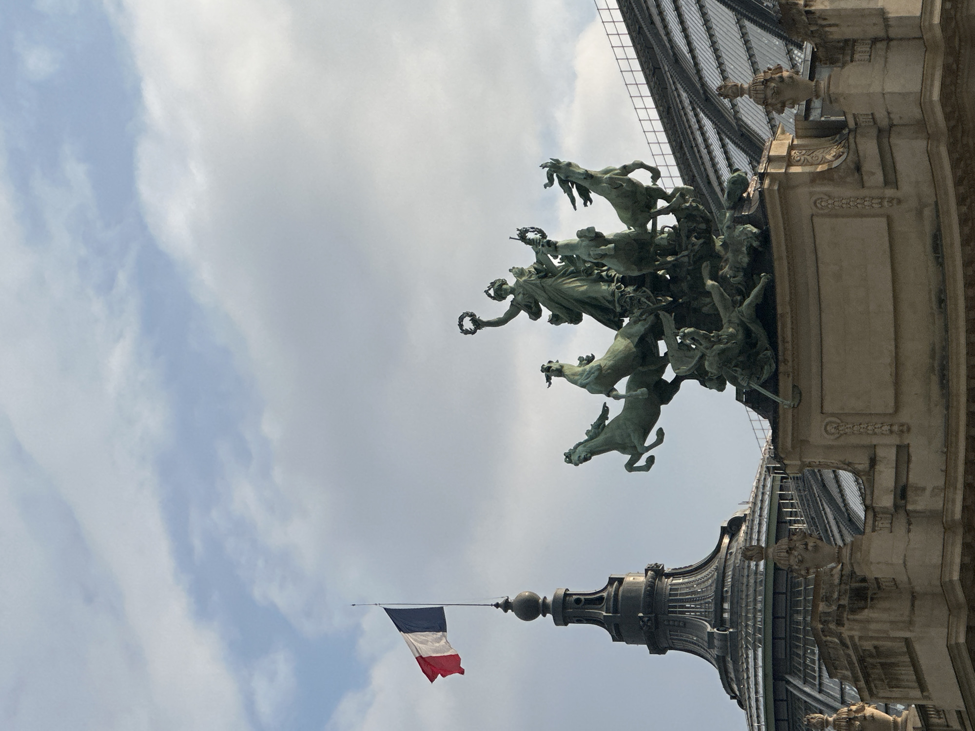 The image depicts a low-angle view of a Parisian landmark. The sculpture of a bronze horse-drawn chariot dominates the upper part of the frame. The architectural features of the building are ornate, with visible stonework and gargoyle-like decorations. The French flag is visible on a spire in the lower portion of the image. The sky is cloudy. The image probably shows part of the Grand Palais.