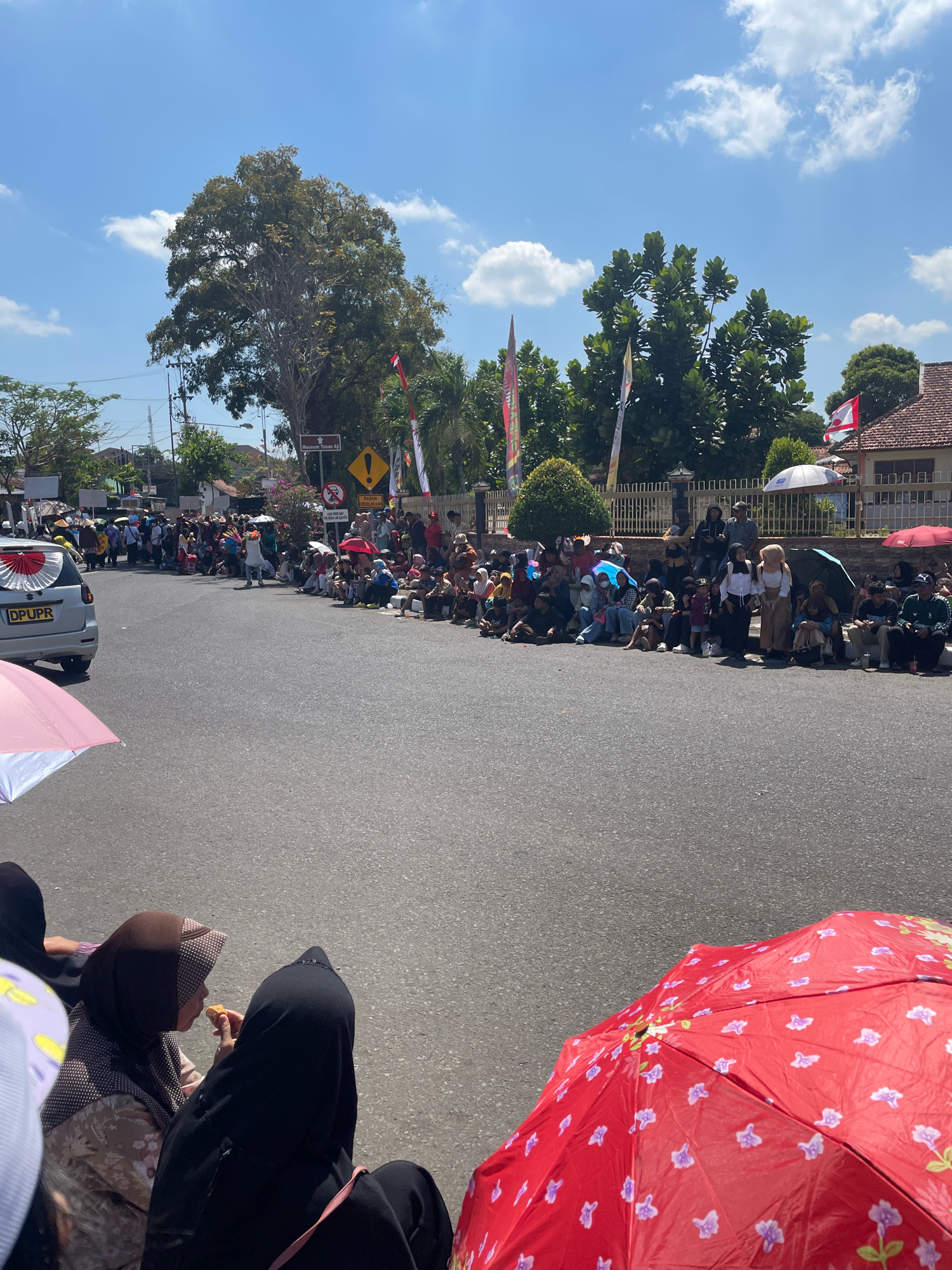 In Purworejo, Indonesia, on a sunny day, a crowd of people is gathered along the side of a street, seemingly watching a parade or event. The people are sitting and standing, some holding umbrellas for shade, including a large red umbrella with a floral pattern in the foreground. A silver vehicle with a license plate that reads "DPUPR" is parked on the left side of the road. In the background, trees, buildings, and flags are visible, suggesting an urban or suburban setting. Some individuals are seen interacting, with one person appearing to eat. The overall atmosphere seems festive and communal.