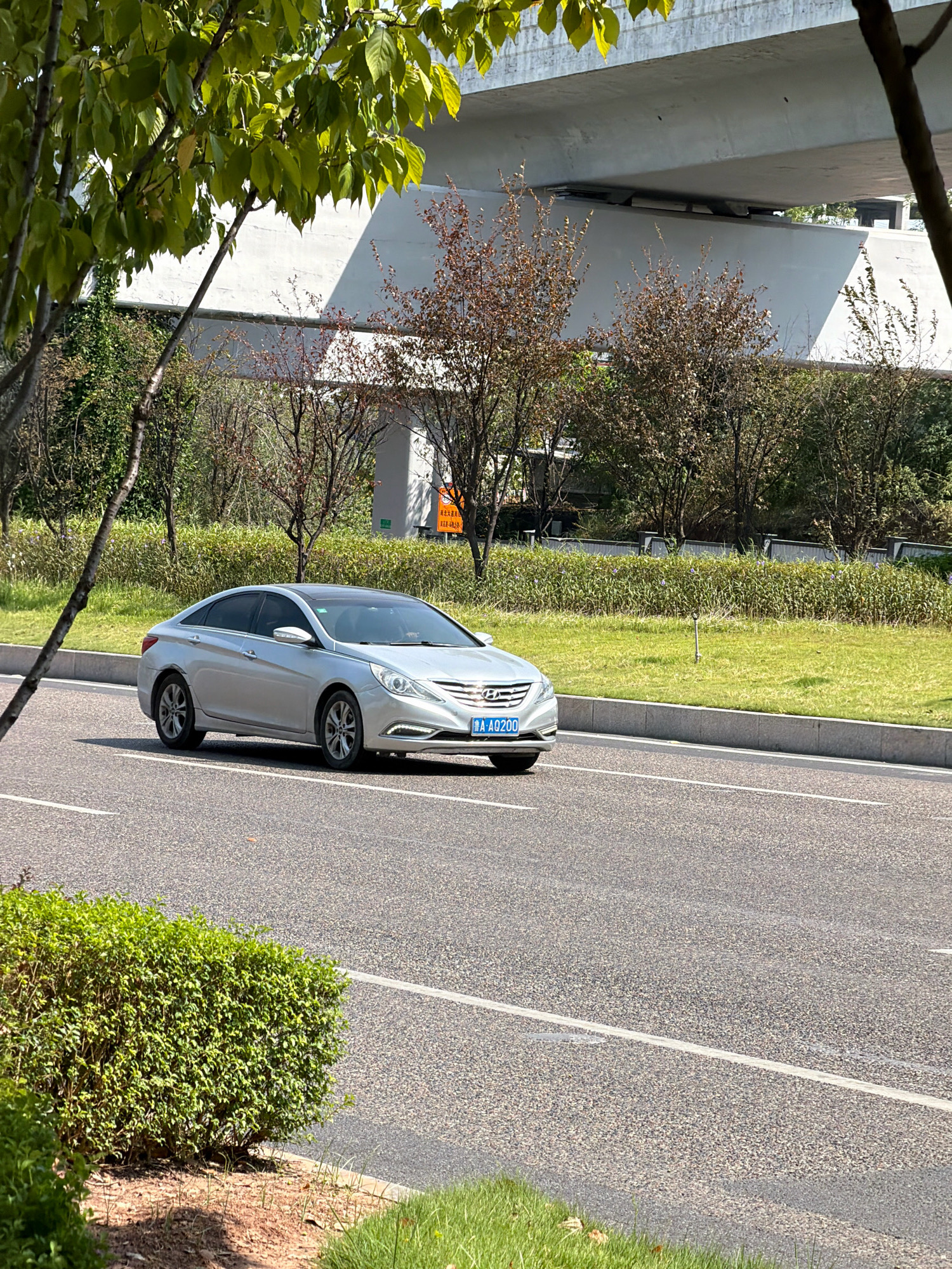 A silver sedan is traveling on a road in Jiangjin, China. The car is a Hyundai Sonata and is driving on the right side of the road. The car has a blue license plate with white characters that read "A AO200". The road is asphalt and has white lane markings. There is a grassy median on the right side of the road, with some trees and bushes. In the background, there is an elevated highway overpass and more trees. The sky is blue and the sun is shining.