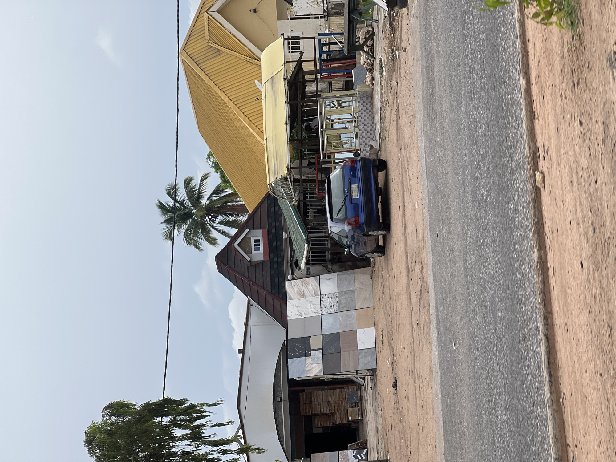 The image depicts an outdoor scene in Jalingo, Nigeria, featuring a small business that appears to sell tiles and possibly construction materials. In the foreground, a paved road runs horizontally, bordered by sandy earth. Behind the road, a blue sedan is parked facing left. To the right of the car, a display rack holds an assortment of square tiles in various patterns and colors, including marble-look, grey, and beige tones. Further back, a building with a distinct gabled roof and a dormer window is visible, along with another structure with a bright yellow, corrugated metal roof. Several people are partially visible within a covered area under the yellow roof, suggesting ongoing business activity. Stacks of boxed materials, likely tiles or construction supplies, are arranged in an open-fronted area to the left. The environment appears to be overcast, with a few trees, including a palm tree, scattered throughout the scene. The overall impression is of a commercial area on a cloudy day.