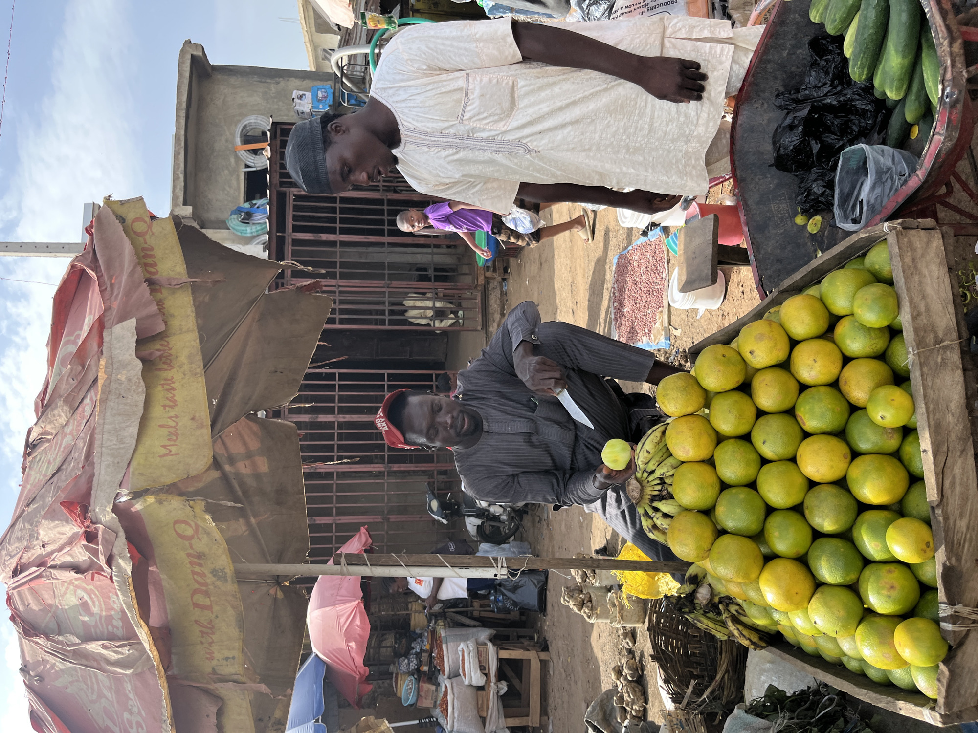 A medium shot captures an outdoor market scene in Rafin Par, Nigeria, under a tattered, faded red and yellow umbrella. Two men are the primary subjects. In the foreground, a man wearing a grey, striped tunic and a red cap is seated, holding a knife in his right hand and a yellow fruit in his left, seemingly preparing to cut it. Before him is a large, wooden crate overflowing with a pyramid of ripe oranges, their skins a mix of green and yellow. To the right of the crate, a portion of a red wheelbarrow is visible, containing black plastic bags and several long, green cucumbers.
Behind the seated man, another man stands, dressed in a cream-colored, loose-fitting tunic with embroidered trim. He faces the seated man, looking downwards. Further in the background, through a metal gate, a child in a purple shirt walks away from the camera, and other market stalls and buildings are dimly visible. The ground is dusty and uneven. The overall impression is that of a vibrant, active marketplace.