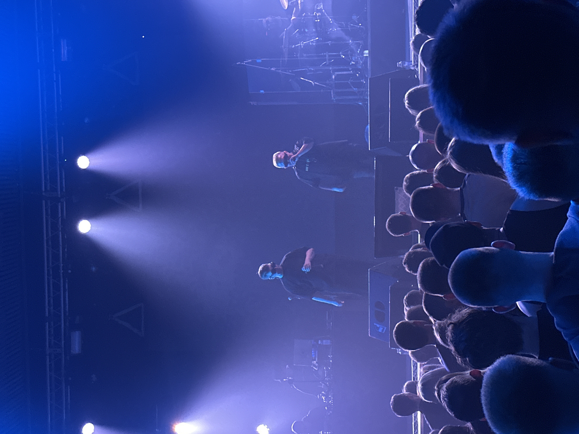 Here is a detailed description of the image:
The image captures a concert scene, likely a musical performance, in a dark venue in Izhevsk, Russia. The foreground is filled with a dense crowd of people, with most of their backs facing the camera. The heads of the audience are mostly visible, with some individuals wearing caps or hoodies. The scene is dominated by blue lighting and stage lights, creating a dynamic and dramatic atmosphere.
On stage, two performers can be seen standing in front of a stage speaker. The performers are in dark clothing. Visible in the background is stage equipment including a drum set and rigging.