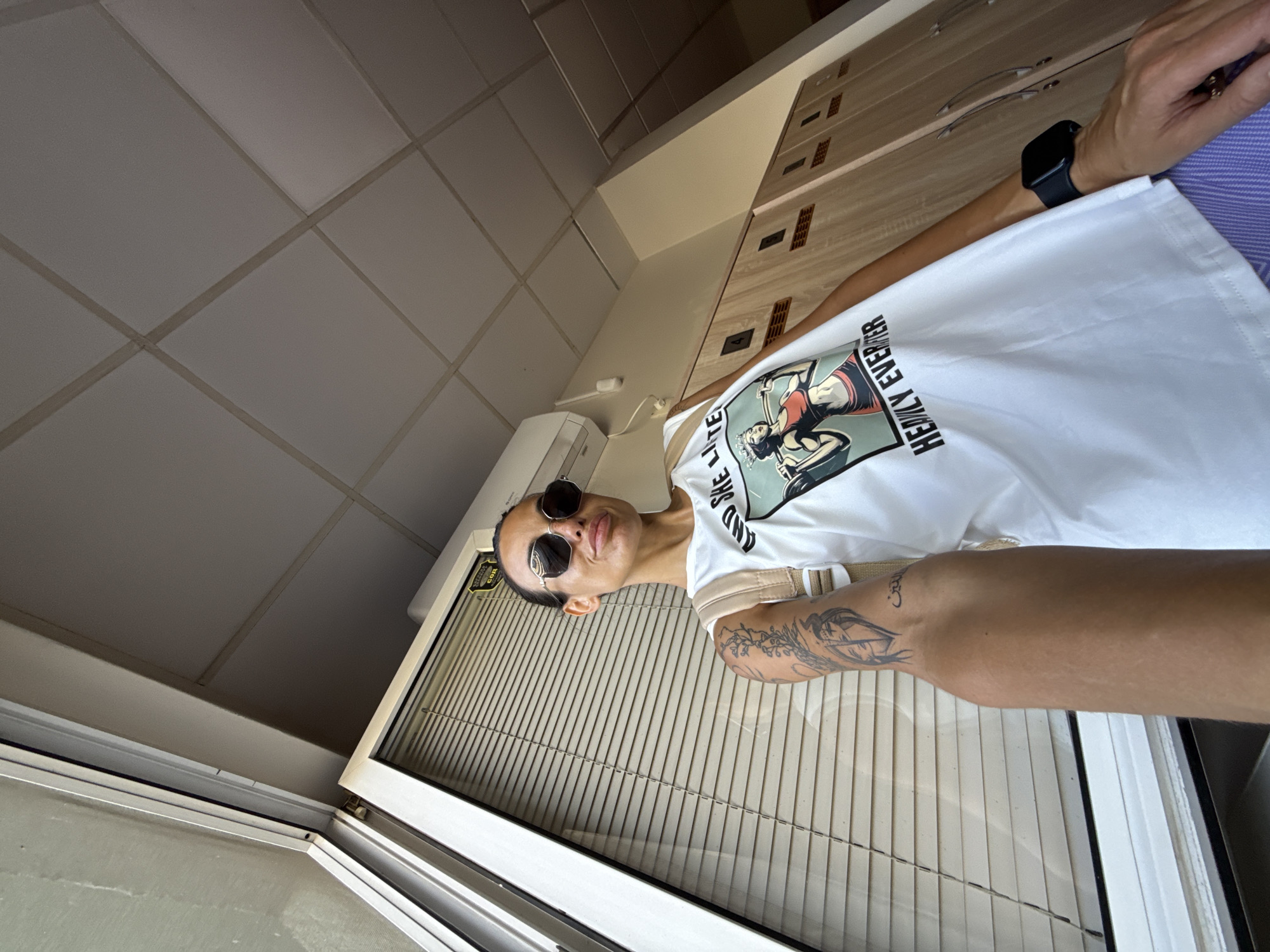 A woman is taking a selfie in a locker room. She is wearing a white t-shirt with a graphic print, sunglasses, and a backpack. She has tattoos on her left arm. The locker room has wooden lockers and a tiled ceiling. The image is taken from a low angle, looking up at the woman.