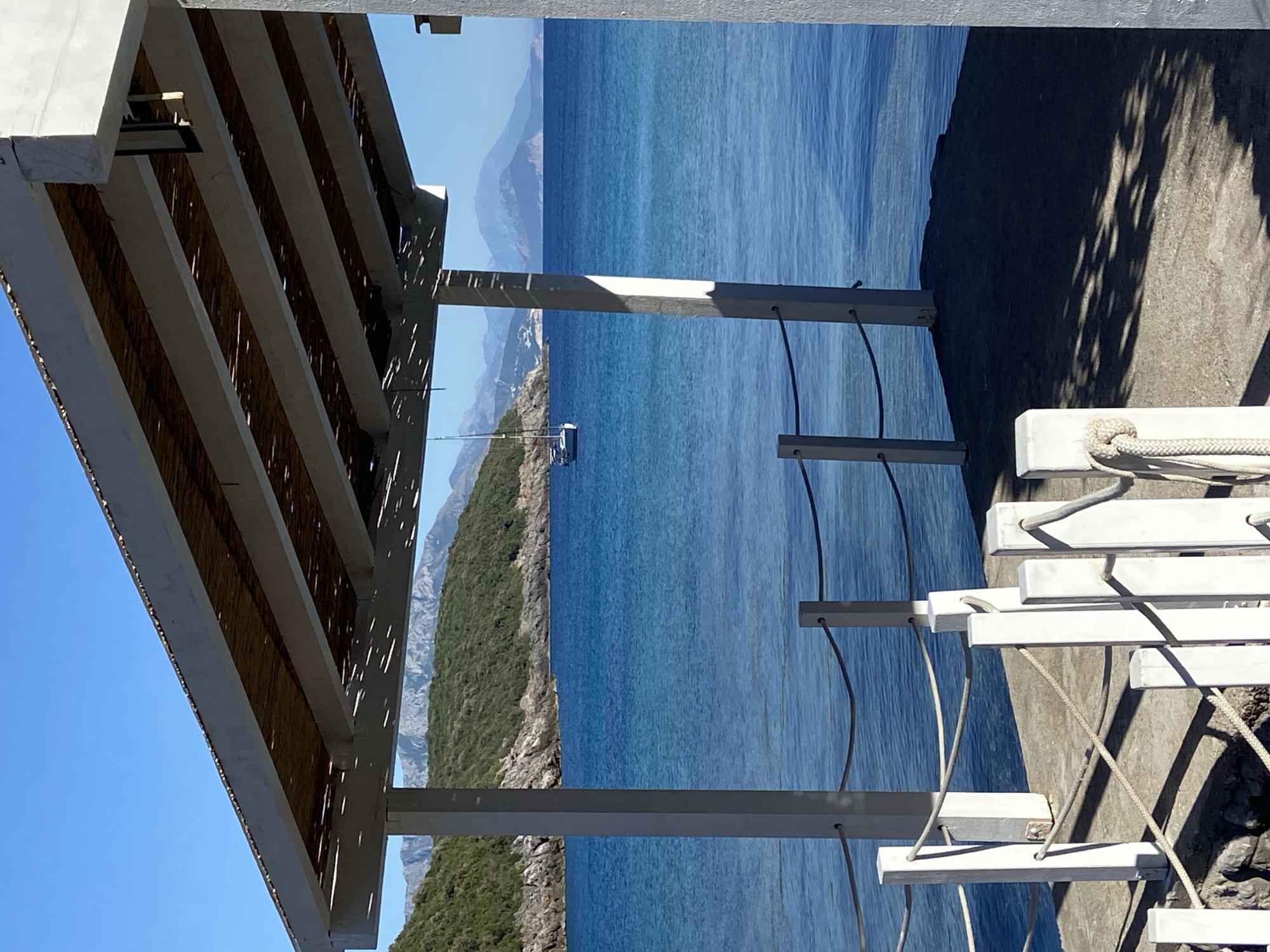The image presents a serene coastal view, likely from a shaded outdoor area, overlooking the sea and a mountainous coastline. No people are visible.
In the upper part of the frame, a white overhead structure, resembling a pergola or balcony ceiling, is prominent. It features parallel concrete beams with a slatted covering, possibly bamboo or reeds, creating patterns of dappled sunlight and shadow.
The mid-ground is dominated by the calm, deep blue expanse of the sea, reflecting the bright sky. In the distance, a small white boat or yacht is seen traversing the water, positioned closer to the right coastline.
To the right, a verdant, rocky coastline stretches along the horizon, leading to several layers of hazy blue mountains under a clear, bright sky. The vegetation appears dense and green.
In the lower right corner, a unique structure resembling a set of white wooden steps or platforms is visible, secured by ropes. These steps descend along what appears to be a concrete or rocky embankment, suggesting access to a lower level or the water's edge. Strong shadows are cast on this surface, indicating bright sunlight.
The scene depicts a sunny, clear day, likely midday or early afternoon, given the intensity of the light and the distinct shadows. The overall atmosphere is one of tranquility and natural beauty, characteristic of a coastal retreat in a location such as Visnjeva, Montenegro. There is no visible text.