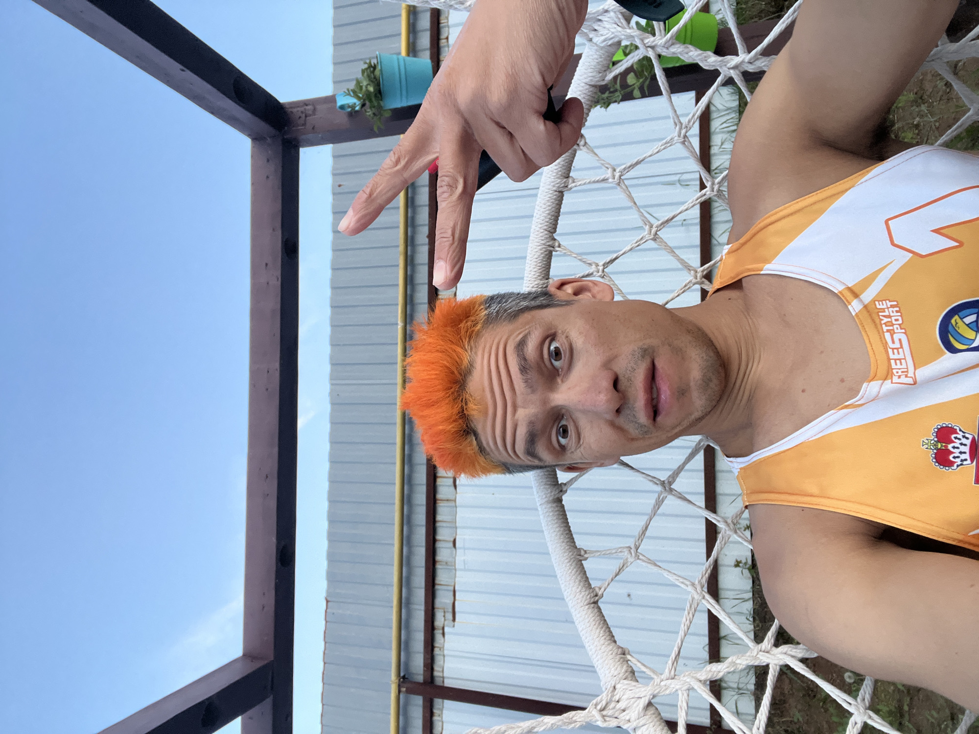 A man with bright orange, spiky hair sits in a white rope swing chair, looking directly at the camera with wide, surprised eyes. He is wearing an orange and white sleeveless athletic shirt. To the right of his head, a hand makes a peace sign gesture. Behind him, a corrugated metal fence runs horizontally, with a wooden structure above it and a clear blue sky beyond. A small blue pot with a green plant is visible to the right of the frame. The location is the city of Chaikovsky, Russia.