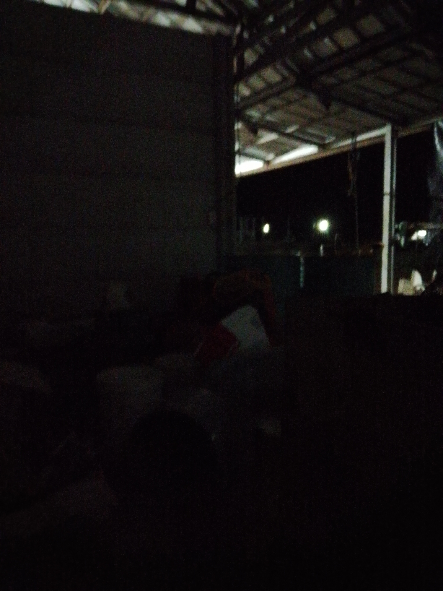 The image, taken at night in Bukit Ketok, Indonesia, shows a dimly lit shed or storage area. Inside, there are various items piled together, making it difficult to discern specific objects. The roof structure is visible, suggesting an open-air or partially enclosed space. Outside, there are lights and indistinct shapes, adding depth to the scene. The overall impression is one of cluttered storage in a low-light environment.