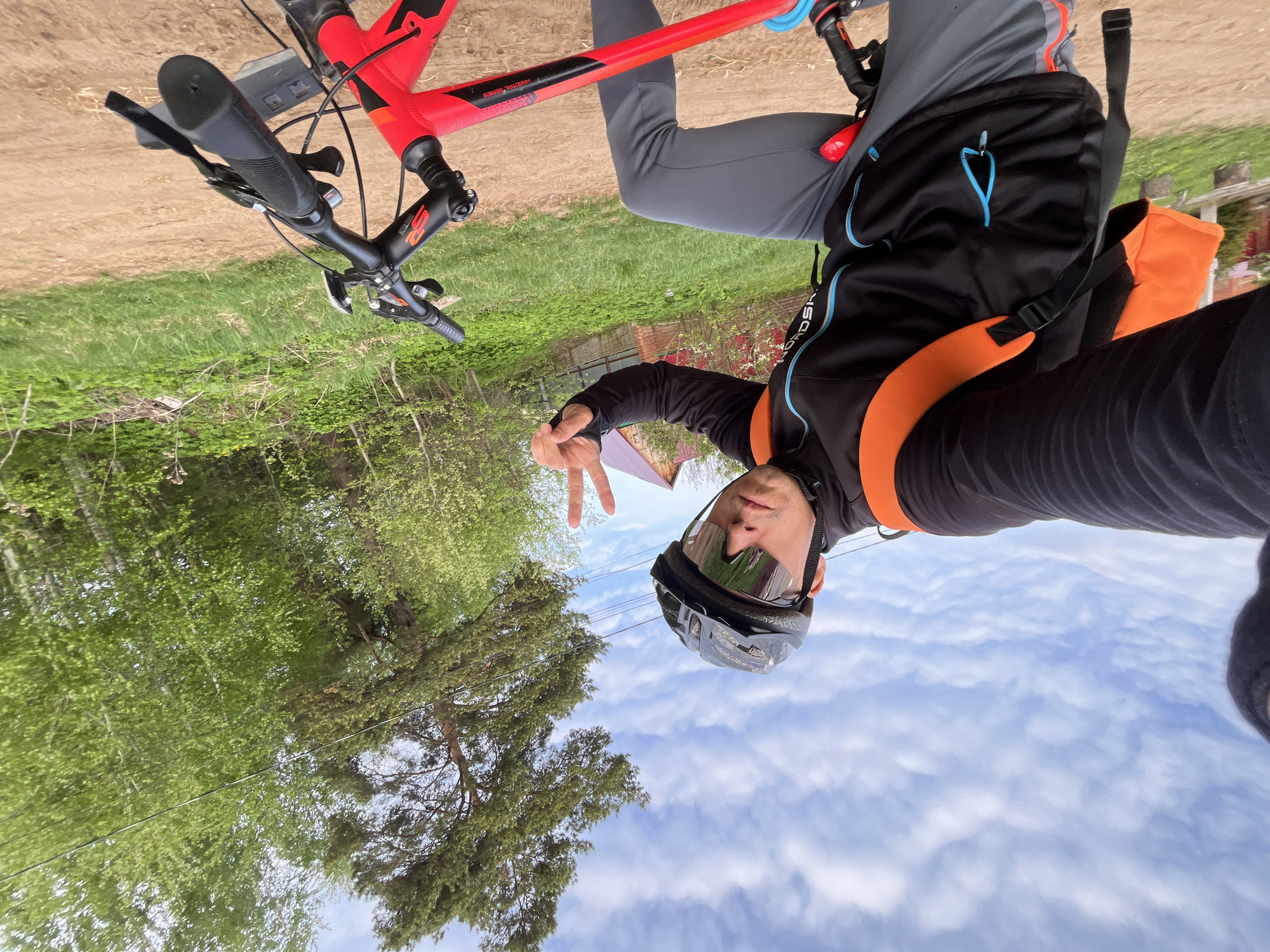 A selfie shows a person wearing a helmet and black and orange jacket, holding up two fingers while riding a red bicycle. The location appears to be in a wooded area, with trees lining a dirt path. The sky is visible with scattered clouds. The city of Chaikovsky, Russia cannot be confirmed from this image.