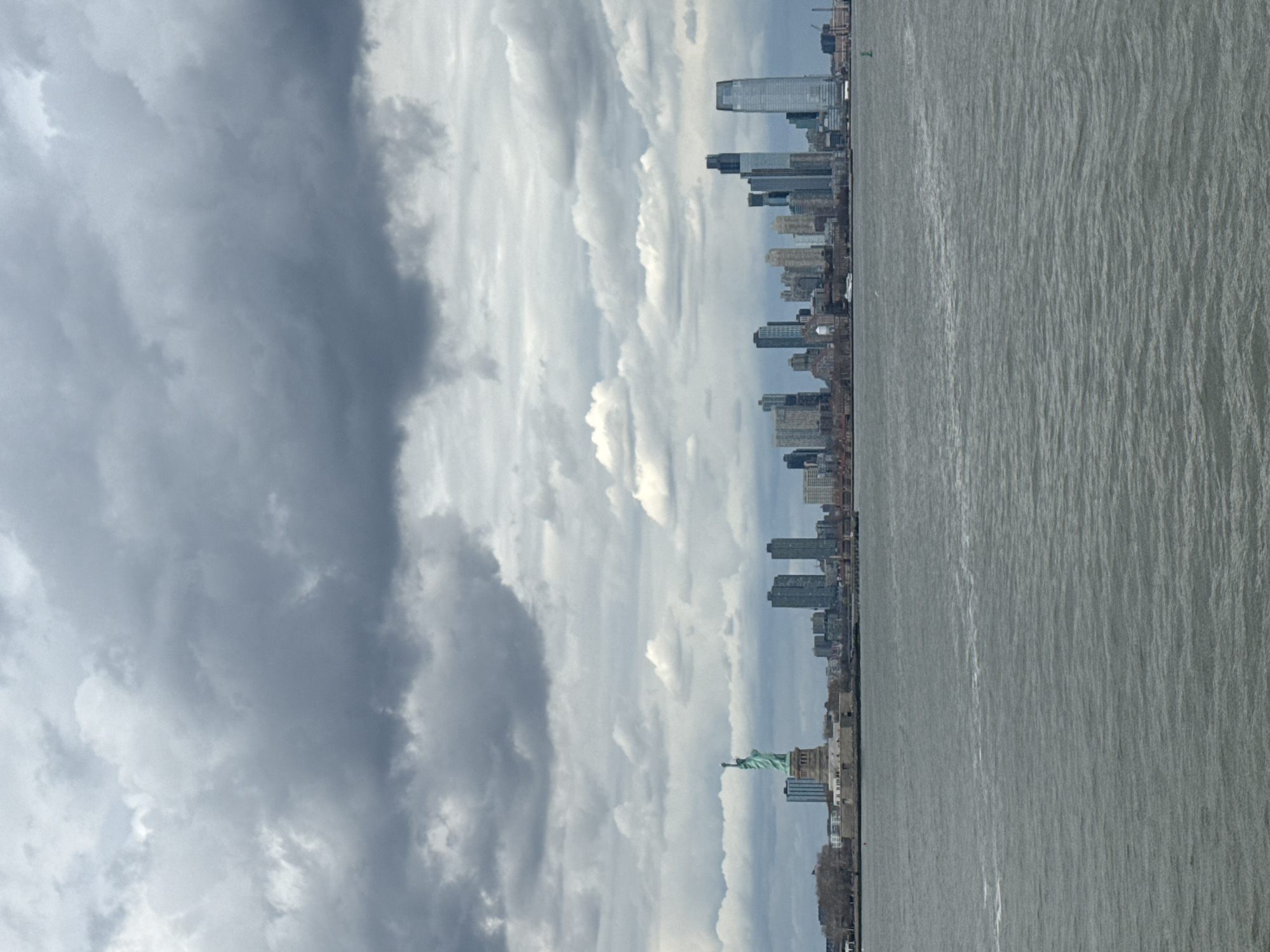 The image shows a cloudy sky above the New York City skyline as viewed from Jersey City, USA. The Hudson River is in the foreground. The Statue of Liberty is also visible in the foreground. The general weather condition are cloudy.