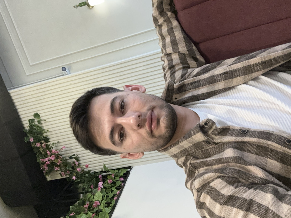 A man with short dark hair and a trimmed beard is angled in the frame, wearing a white waffle-textured shirt and an unbuttoned, striped flannel. He is seated, with the back of a burgundy chair visible. Behind him is a white, slatted wall with flowers and greenery in the lower left corner. A light fixture and a security camera are seen on the ceiling. The photograph appears to have been taken indoors.