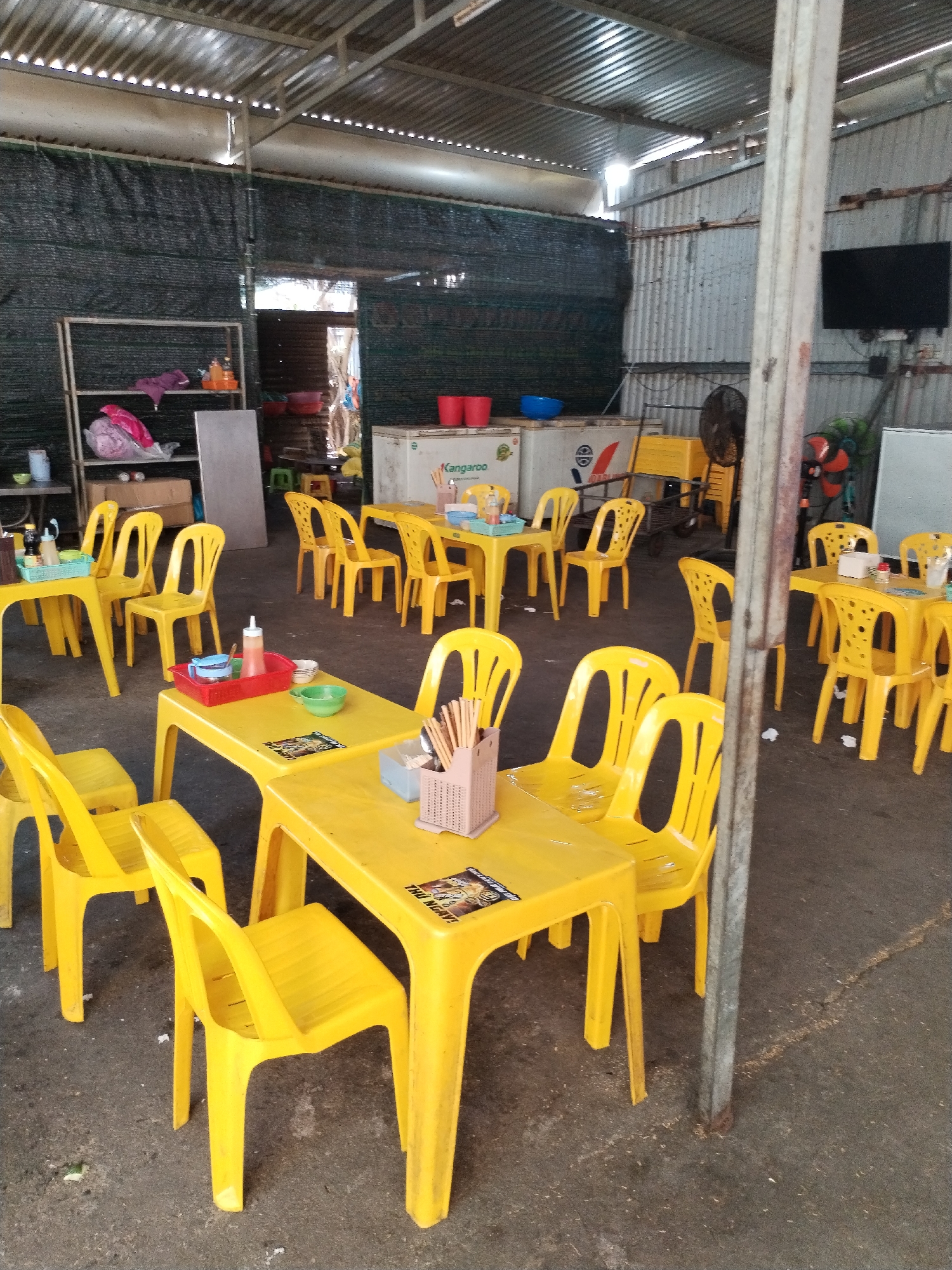 This image captures an outdoor dining space in Nha Trang Ward, Vietnam. The setting is characterized by yellow plastic tables and chairs. The tables are set with condiments, chopsticks, and napkins. The flooring is concrete with visible staining. In the background are metallic walls, a storage rack, and a cooler with the word "Kangaroo" on it. A television is mounted on the metallic wall. No people are visible.