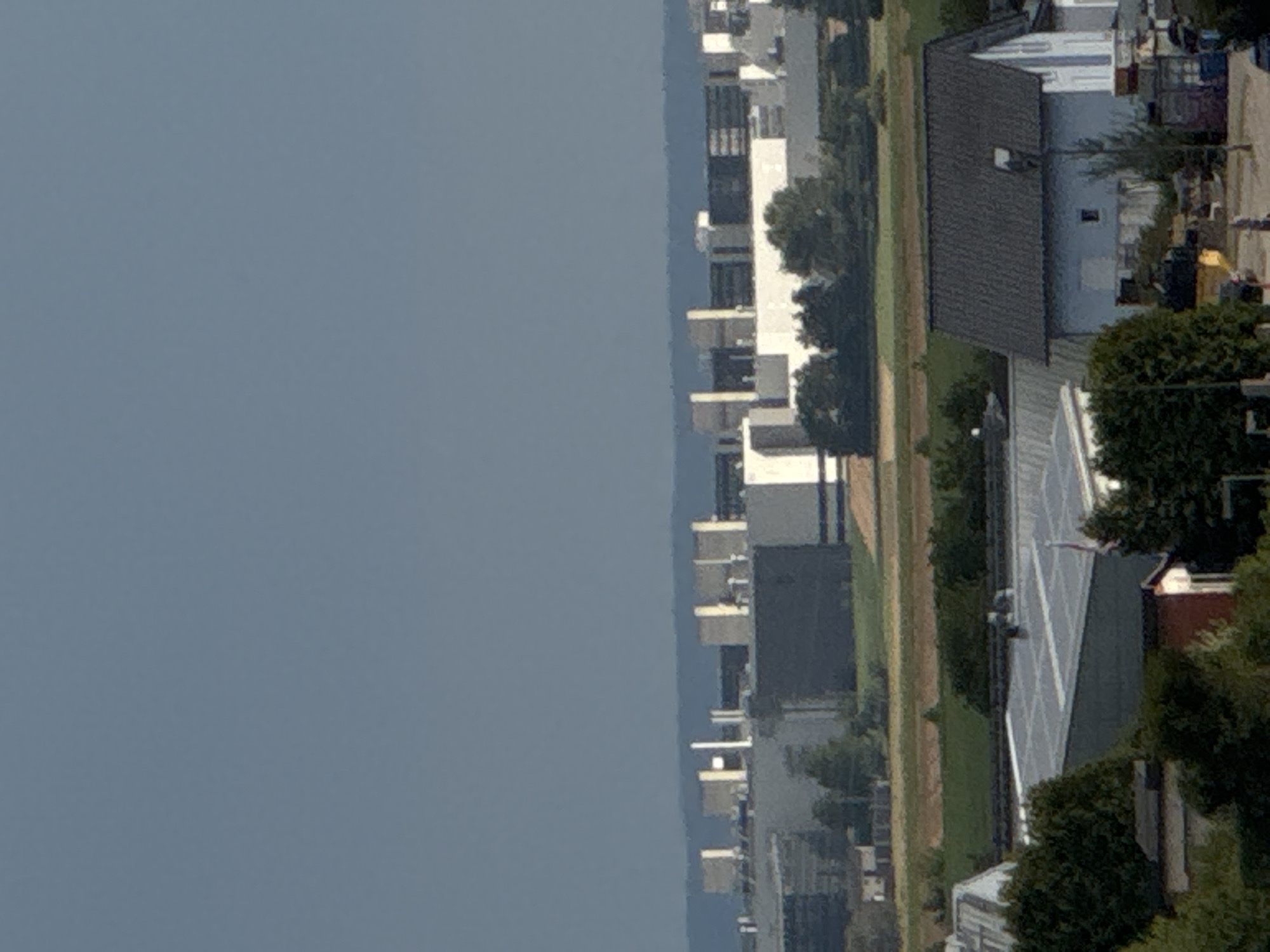 The image shows a panoramic view of Wettstetten, Germany, featuring a mix of industrial buildings and residential structures. In the background, a large, gray industrial complex with numerous rectangular sections and white vertical elements dominates the horizon. The sky above is a clear, pale blue. In the middle ground, a green field separates the industrial area from a collection of residential buildings. These buildings include a light blue house with a dark gray, gabled roof and a small window on the upper floor, as well as other structures with lighter colored roofs. Trees are scattered throughout the scene, particularly around the residential areas. In the foreground, more trees and some smaller buildings are visible, including one with a red roof and another with a white, sloped roof. The overall impression is of a semi-rural industrial town.