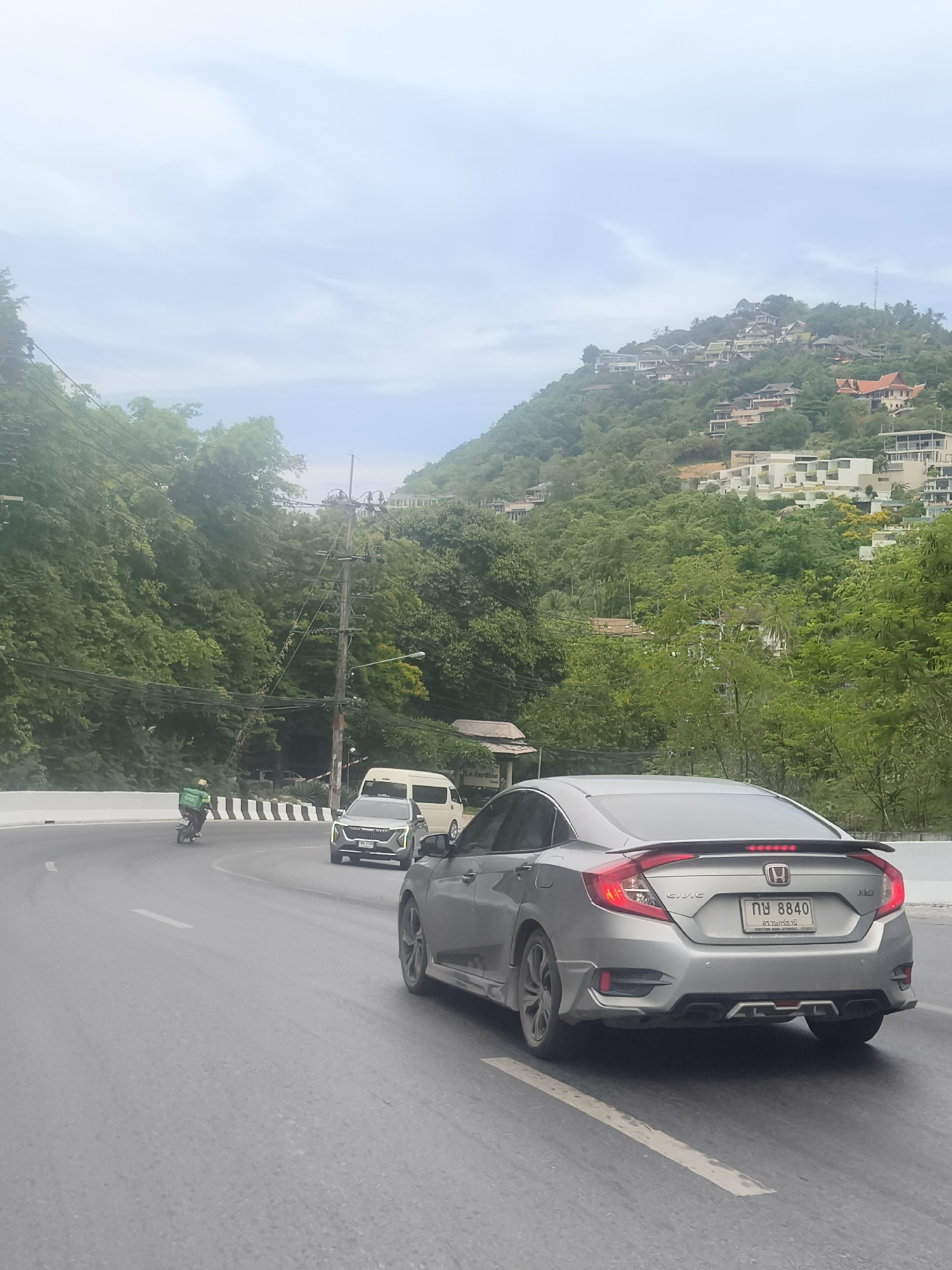 An overcast day frames a scenic road curving through a lush, green hillside in Baan Chaweng Noi, Thailand. Multiple houses are visible nestled on the slope. A silver Honda Civic sedan, with license plate 'กญ 8840' and 'สยามธานี' visible, is traveling on the right side of the road. A grey SUV is in front of it, and a white van is further ahead. A motorcyclist wearing a green jacket with a 'Grab' logo is on the left side of the road, appearing to be making a delivery. Power lines crisscross overhead, supported by poles along the roadside, and dense foliage covers the surrounding hills.