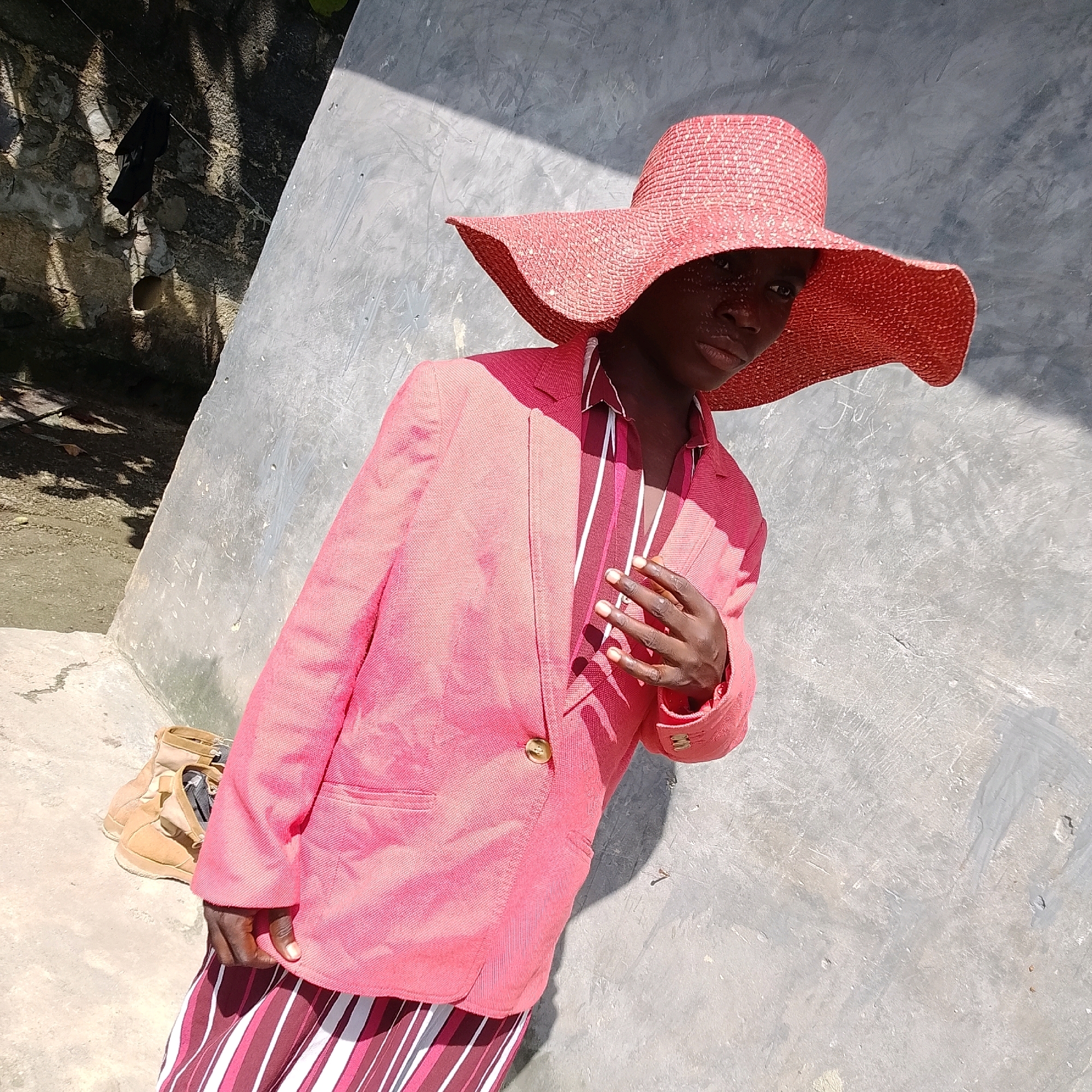 A person stands outside near a concrete wall, leaning towards the camera. The person is wearing a wide-brimmed red hat, a pink blazer with gold buttons, and a striped skirt in shades of red, white, and dark pink. They are also wearing a shirt with thin vertical stripes. A pair of tan boots are visible on the ground to the left of the person. Sunlight casts shadows on the person and the surrounding environment, which appears to be an outdoor area with a rough stone wall on the left and a textured concrete wall on the right. The ground is a mix of concrete and dirt. The scene is likely in Ado, Nigeria, suggested by the context provided.