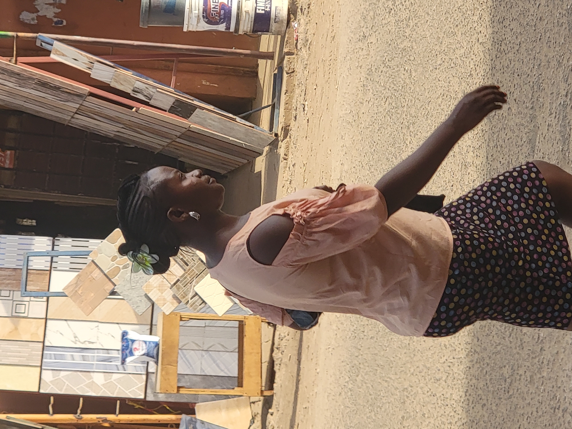 A woman in a pale pink blouse and a dark, polka-dotted skirt stands outside of a shop in Maiburiji, Nigeria. Her hair is braided, and she wears a white flower tucked behind her ear. Her right arm is extended upward. The shop appears to sell tiles and paint buckets. The ground is light-colored, possibly concrete or pavement. The location is likely a street with storefronts.