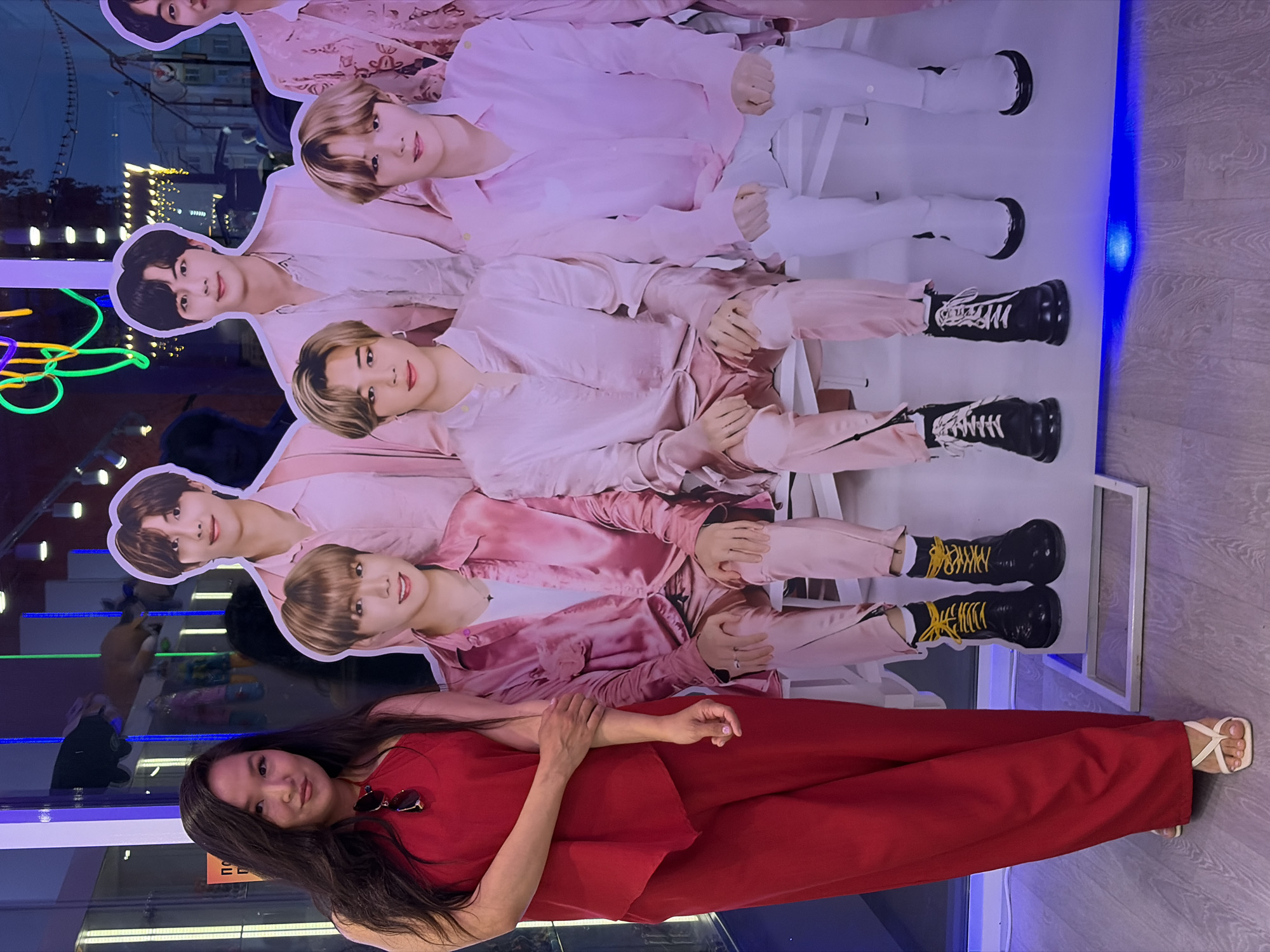 A woman is lying down in front of a large poster of the K-pop band BTS. The poster is vertically oriented, with the band members positioned from top to bottom. The woman is wearing a red dress and white sandals. She is smiling. The location appears to be indoors, possibly a store or entertainment venue in an unknown city. There are reflections of the city visible in the glass windows, including neon lights.