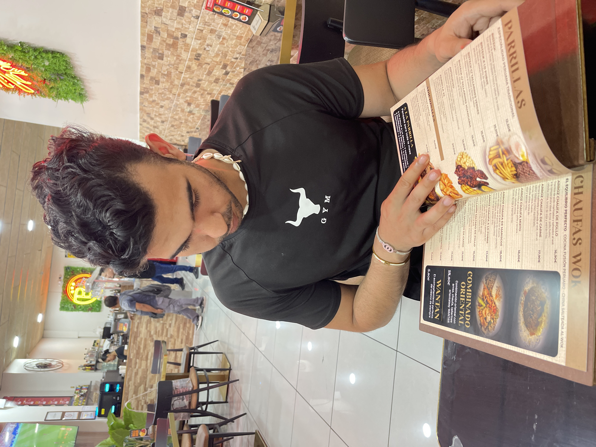 A young man with dark, curly hair and a goatee is sitting at a table in a restaurant, looking down at a menu. He is wearing a black t-shirt with a white bull logo and the word "GYM" underneath it, and a white shell necklace. He is also wearing a gold bracelet with a pink charm. The restaurant appears to be casual, with tiled floors, brick-patterned walls, and bright lighting. In the background, other patrons and staff can be seen. On the wall, there is a neon sign and a television screen showing a soccer game. The menu he is holding is open and displays food items with images. The establishment is located in Santa Coloma de Gramenet, Spain.