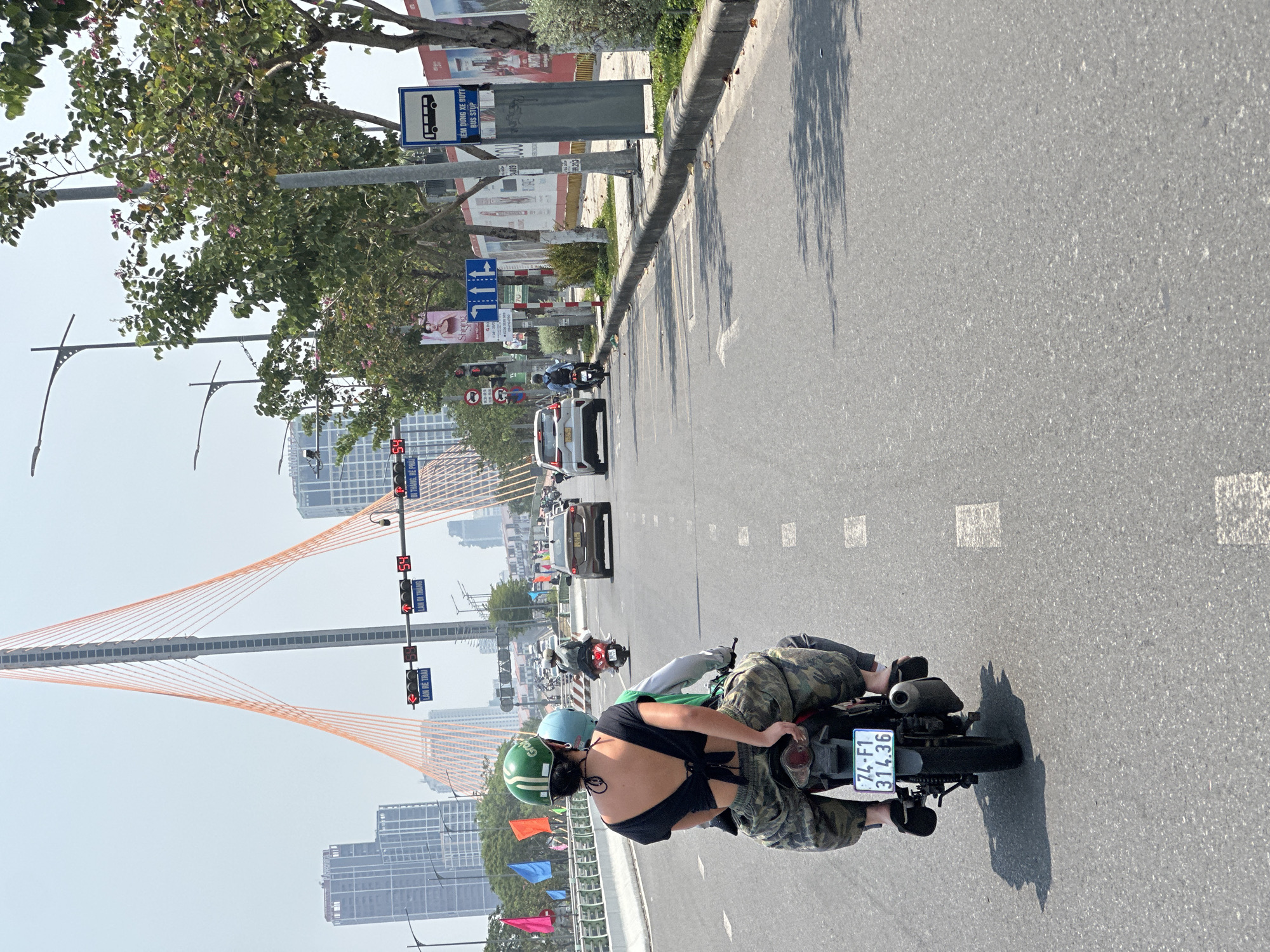 The image shows a street scene in Đà Nẵng, Vietnam, featuring a two-lane road with white lane markings. A person wearing a black top, camouflage pants, and a green helmet is riding a motorcycle. The license plate number is 74-F1 314.36. In the background, the Trần Thị Lý Bridge is visible, along with buildings, traffic lights, road signs (including a bus stop sign), and other vehicles. The weather appears to be clear and sunny.