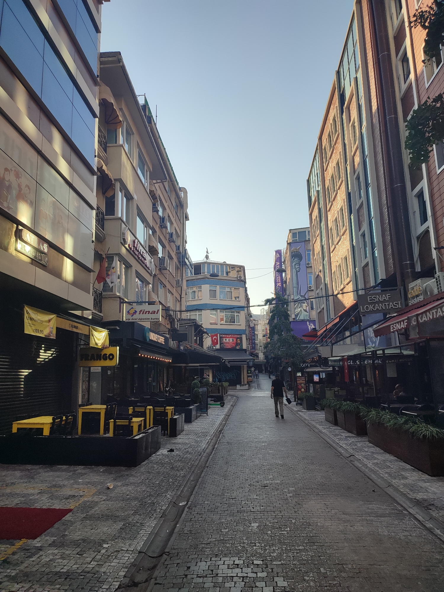 In the city of Kadıköy, Turkey, a cobblestone street is lined with multi-story buildings. The buildings feature a mix of modern and older architectural styles, with many windows and some businesses with awnings. On the left side of the street, yellow tables and chairs are set up outside what appears to be a restaurant or cafe. Several signs with text are visible on the buildings, including one that reads "ESNAF LOKANTASI." A single person, wearing a dark shirt and light-colored pants, walks away from the camera down the middle of the street. The sky above is clear and blue. The scene is a street view with commercial establishments and pedestrian activity.