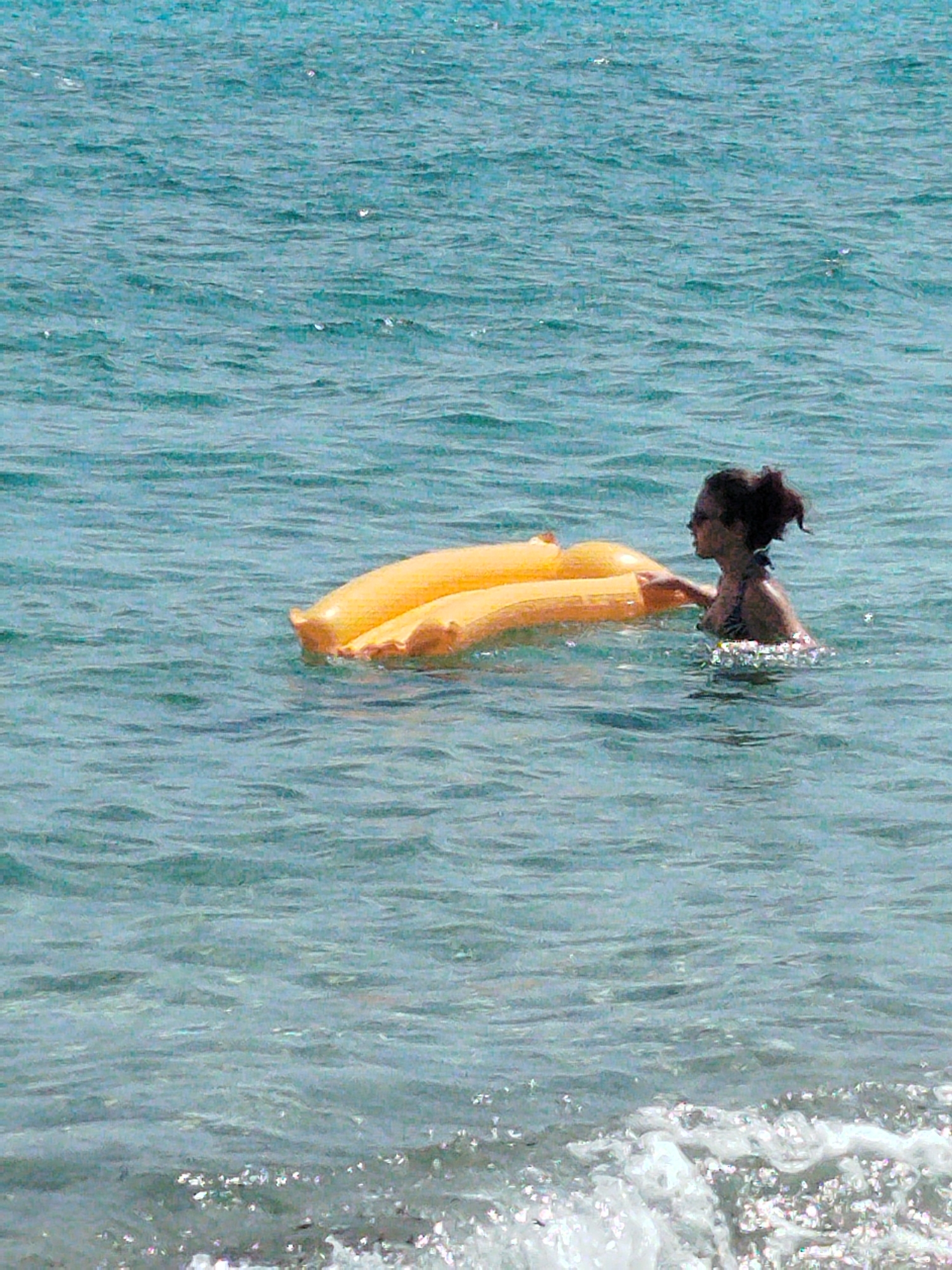 A woman with dark hair pulled back is swimming in the ocean. She is wearing a dark bikini and has her right hand on a large, inflatable yellow raft. The water is a light blue-green color with small waves. The scene appears to be a sunny day, likely in a coastal area such as Sithonia Municipal Unit, Greece.