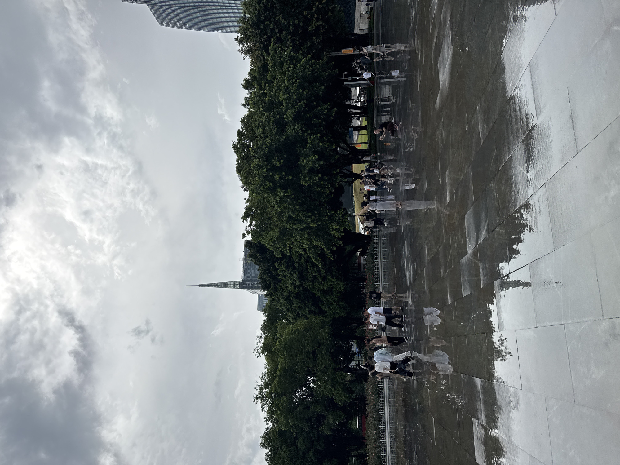 A wide shot captures a wet, reflective plaza in Tianhe District, China, under an overcast sky. The plaza is paved with large, dark gray tiles that mirror the sky, trees, and people. In the mid-ground, a line of lush green trees forms a dense canopy. Behind the trees, a tall, slender tower with a pointed spire rises above the cityscape, with a modern skyscraper visible to the right. Several groups of people are scattered across the plaza, some walking, others standing or crouching. A woman in a light-colored dress stands with her arm raised, possibly posing for a photo. A man is crouched down, appearing to take a picture with his phone. The overall impression is a public space after a rain shower, with people enjoying the cool, damp atmosphere.