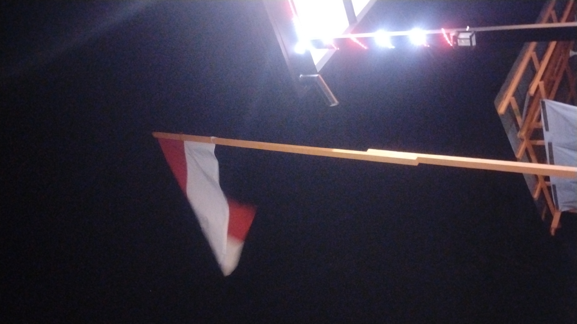 The image shows a flag that appears to be the Indonesian flag, with red on top and white on the bottom, flying from a wooden pole against a dark sky. To the right of the flag, there is a lit structure with several lights, some of which appear to be red. Below the flag and lights, a wooden roof structure is visible. The scene is dark, suggesting it is nighttime. The setting appears to be outdoors, possibly in a public area. The provided information suggests this scene is located in the city of Samarinda, Indonesia. There are no people visible in the image.