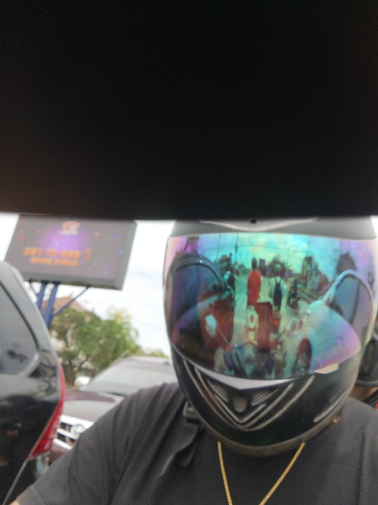 The image is a selfie taken from the perspective of someone wearing a motorcycle helmet. The helmet has a reflective visor that shows a distorted view of the surrounding environment, including other vehicles and people on what appears to be a street. In the background, a digital billboard is visible with some text and graphics, which can be identified as being located in Denpasar, Indonesia due to the text "Denpasar" and a phone number format common in Indonesia. The billboard displays the text "GET TO BBD" and "SPADE HATILE", suggesting it might be an advertisement. The scene appears to be an urban setting, possibly a traffic intersection given the presence of multiple vehicles. The lighting suggests it is daytime.
