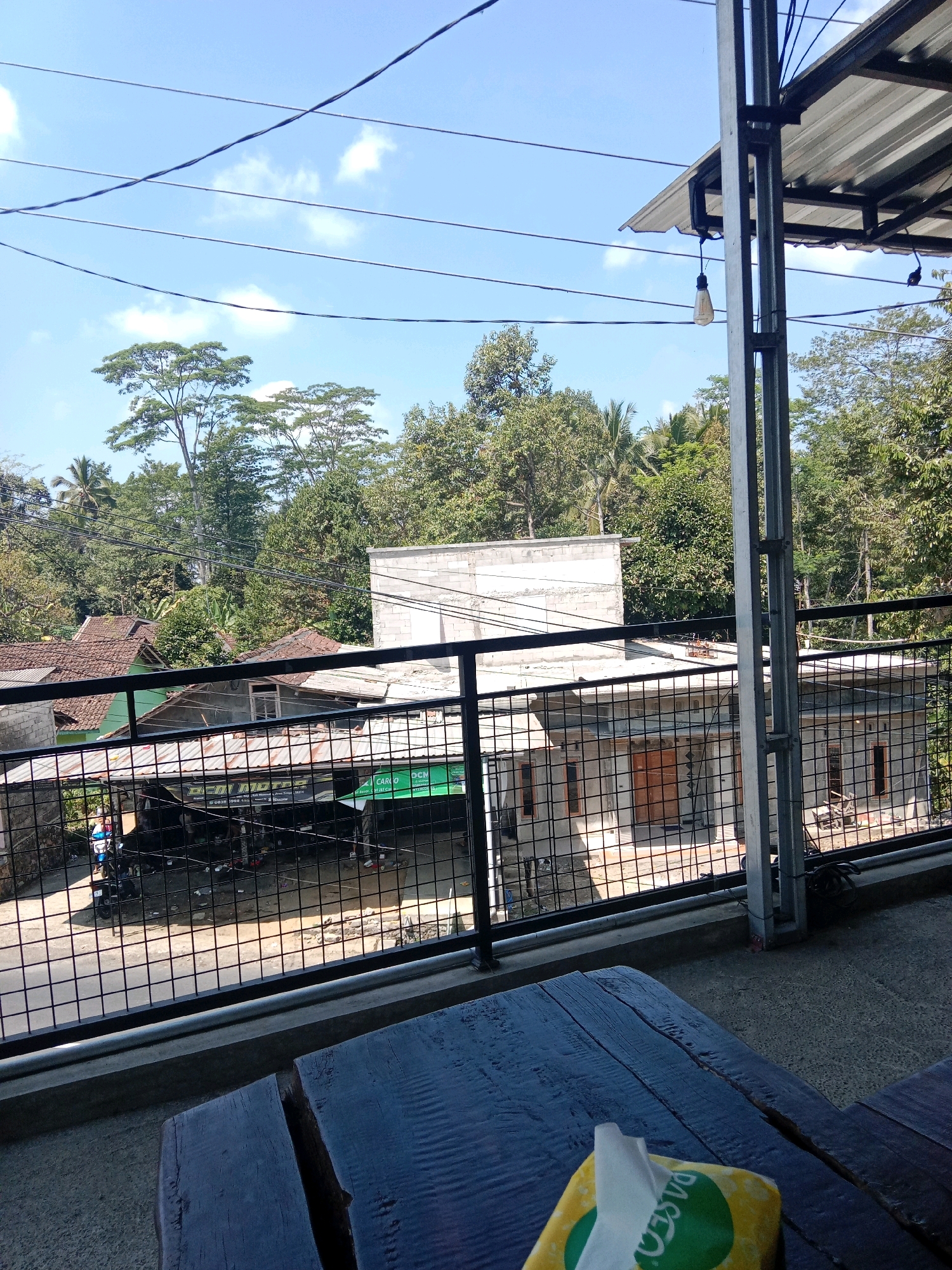 A high-angle, full shot captures a view from a covered patio area overlooking a street in Tanjunganom, Indonesia. The foreground features a dark wooden table with a yellow tissue box and a white tissue in focus. Beyond the table, a black metal railing separates the patio from the street below. The street itself is lined with various buildings, including houses with tiled roofs and what appears to be a small business with a metal awning. A motorcycle is parked on the side of the road. Lush green trees and vegetation fill the background, with a clear blue sky dotted with white clouds overhead. A single light bulb hangs from the metal structure supporting the patio roof. The overall scene suggests a casual, outdoor setting in a semi-rural or suburban area.