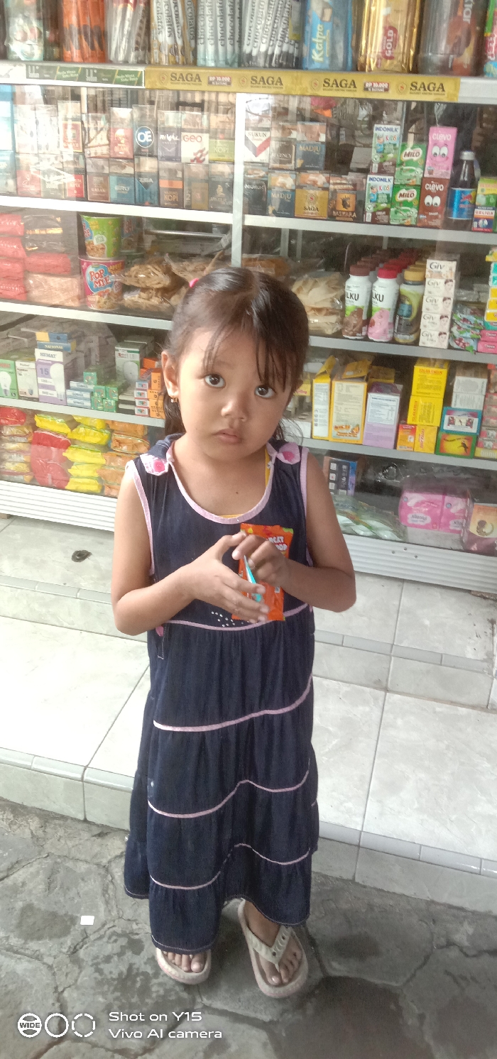 A young East Asian girl is depicted standing in what appears to be the entrance or interior of a small retail shop, likely a warung, in Sebaung, Indonesia. She is positioned centrally, facing directly towards the camera with a neutral expression. The girl has dark hair, bangs over her forehead, and small earrings. She is wearing a navy blue sleeveless dress with pink trim accents and light-colored flip-flops. Her hands are clasped in front of her torso, holding an orange-colored packet, possibly a snack or candy, with a small blue or light green utensil partially inserted into it. The text "GREAT SHOP" is partially visible on the packet.
Behind the girl, a multi-tiered metal shelving unit is visible, displaying a dense array of packaged goods. The top shelves contain numerous cigarette packs, including brands such as SAGA, OE, GEO, SUKUN, KOPi, MADJU, and BASWARA, with "Rp 10.000 / BATANG" pricing visible on yellow banners. The middle and lower shelves are stocked with various consumer products, including instant noodles (Pop Mie, Indomie), bagged snacks, milk and powdered drink boxes (Milo, Indomilk Kids, Clevo), bottled beverages (ULKU), and personal care items (GIV soap, sanitary pads).
The girl is standing on a light-colored tiled step or platform. Immediately in front of this platform, a rough, dark grey concrete or stone surface, possibly a sidewalk or outdoor ground, is visible. The overall scene indicates a typical local convenience store setting.