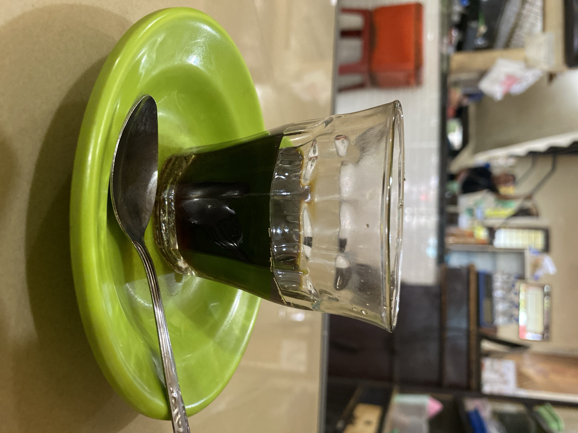 A glass of dark liquid, likely coffee, sits on a bright green saucer. A metal spoon rests on the saucer to the right of the glass. The background is blurred but suggests an indoor setting, possibly a cafe or shop, with shelves and furniture visible. The overall impression is of a simple, everyday scene. While the image itself does not contain enough information to confirm the specific location of Lhokseumawe, Indonesia, the scene is consistent with a local establishment in such a city.