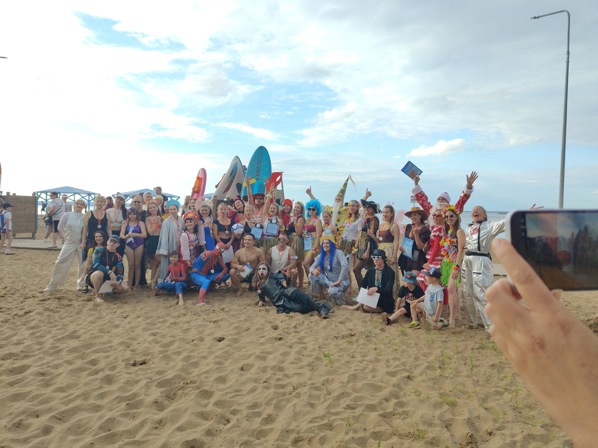 A large group of people in various costumes are gathered on a sandy beach, posing for a photograph. The individuals are wearing a mix of colorful and thematic outfits, including what appears to be a Santa Claus costume, a Spiderman costume, and Hawaiian-themed attire. There are several surfboards and other watercraft standing upright behind the group. The environment is an outdoor beach setting with a cloudy sky overhead. The people are arranged in multiple rows, with some crouching and others standing. The overall atmosphere seems festive and casual.