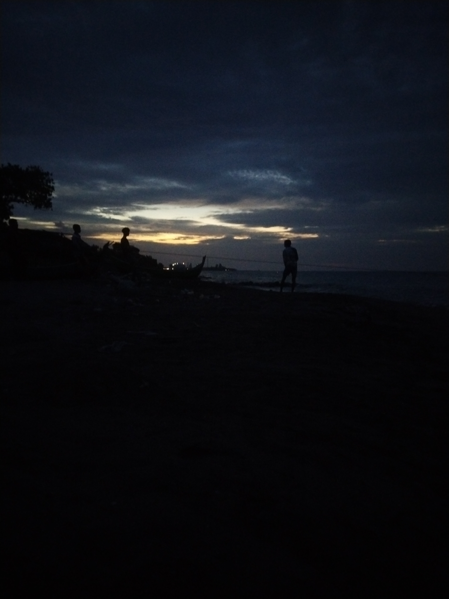 The image captures a dimly lit scene at dusk or dawn on a beach, possibly in Lhokseumawe, Indonesia. The sky is a deep blue with streaks of lighter clouds, suggesting the last vestiges of daylight or the first hints of morning. In the foreground, the dark silhouette of a beach is visible, with a few indistinct figures in the distance. On the left side, a tree is silhouetted against the sky. Further to the right, near the water's edge, several people are seen as dark figures, some appearing to be sitting or standing near a boat. One person stands closer to the water, looking out towards the sea. The water itself is a dark expanse, with a faint horizon line. The overall impression is one of quiet solitude and the natural beauty of a coastal landscape at twilight.