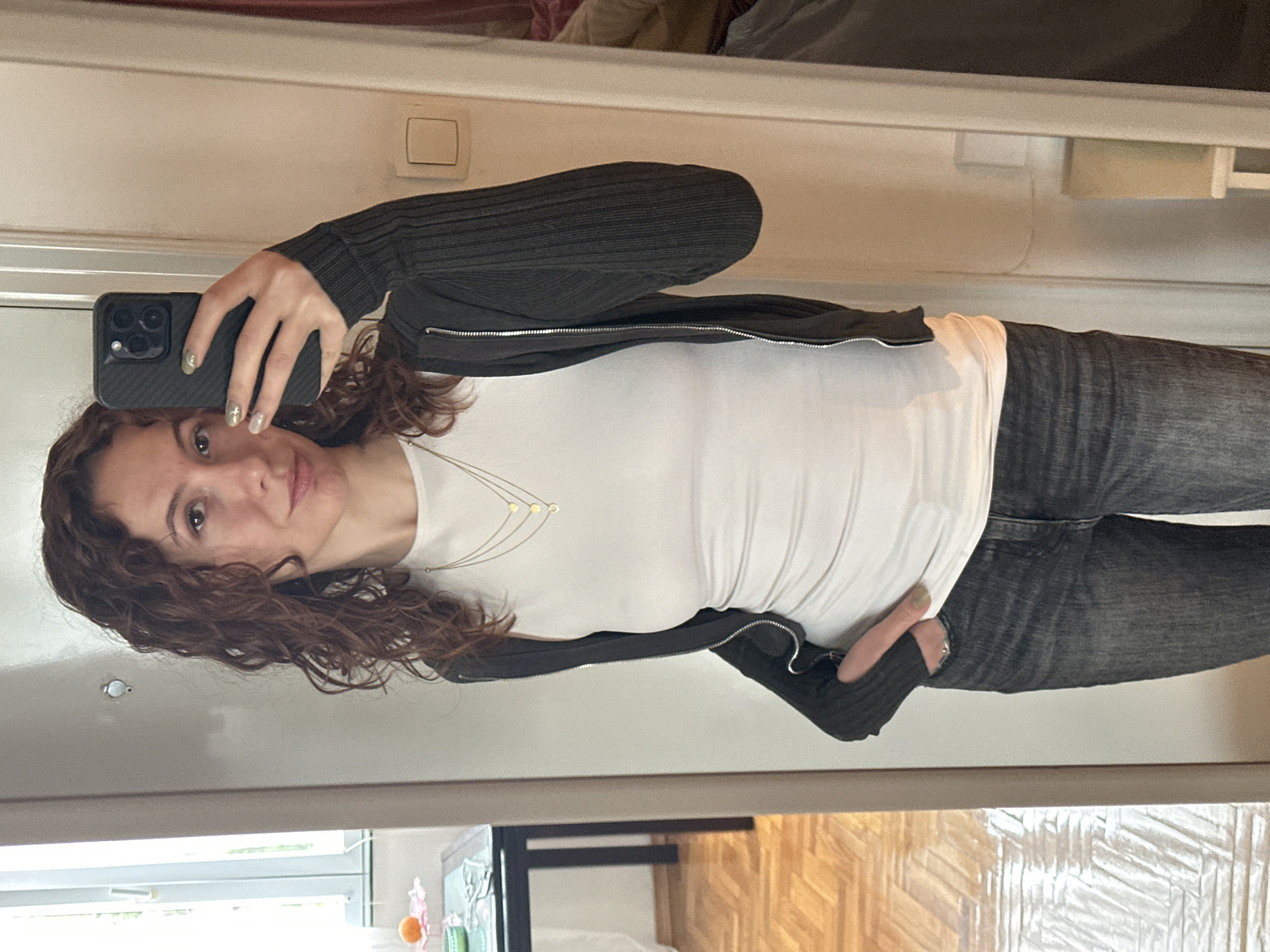 An indoor, vertical mirror selfie captures a young woman from the chest up, slightly angled to the side. She has curly, shoulder-length brown hair and a light complexion, looking towards the camera with a subtle smile. She is holding a dark-colored iPhone with a textured black case in her right hand, which is visible in the upper left of the frame. Her nails are painted a light green or gold shade, some adorned with small star or cross designs.
The woman is wearing a white, fitted long-sleeved top, layered with a dark grey or black ribbed zip-up cardigan or jacket. A delicate gold-colored necklace with small, scattered circular or heart-shaped pendants is visible around her neck. Her left hand is casually placed near her waist, showing a ring on her finger. She is also wearing dark grey jeans.
The background, seen through the mirror's reflection, consists of plain light-colored walls. A white light switch is visible on the wall behind her head. Towards the bottom of the frame, a wooden floor with a parquet or herringbone pattern can be seen. Other blurry shapes suggest typical indoor furniture or decor, including what might be a window frame or another reflective surface further in the background. The lighting suggests an indoor setting, likely during the day, with no strong natural light sources directly visible. No legible text is visible in the image. The general atmosphere conveyed is casual and relaxed.