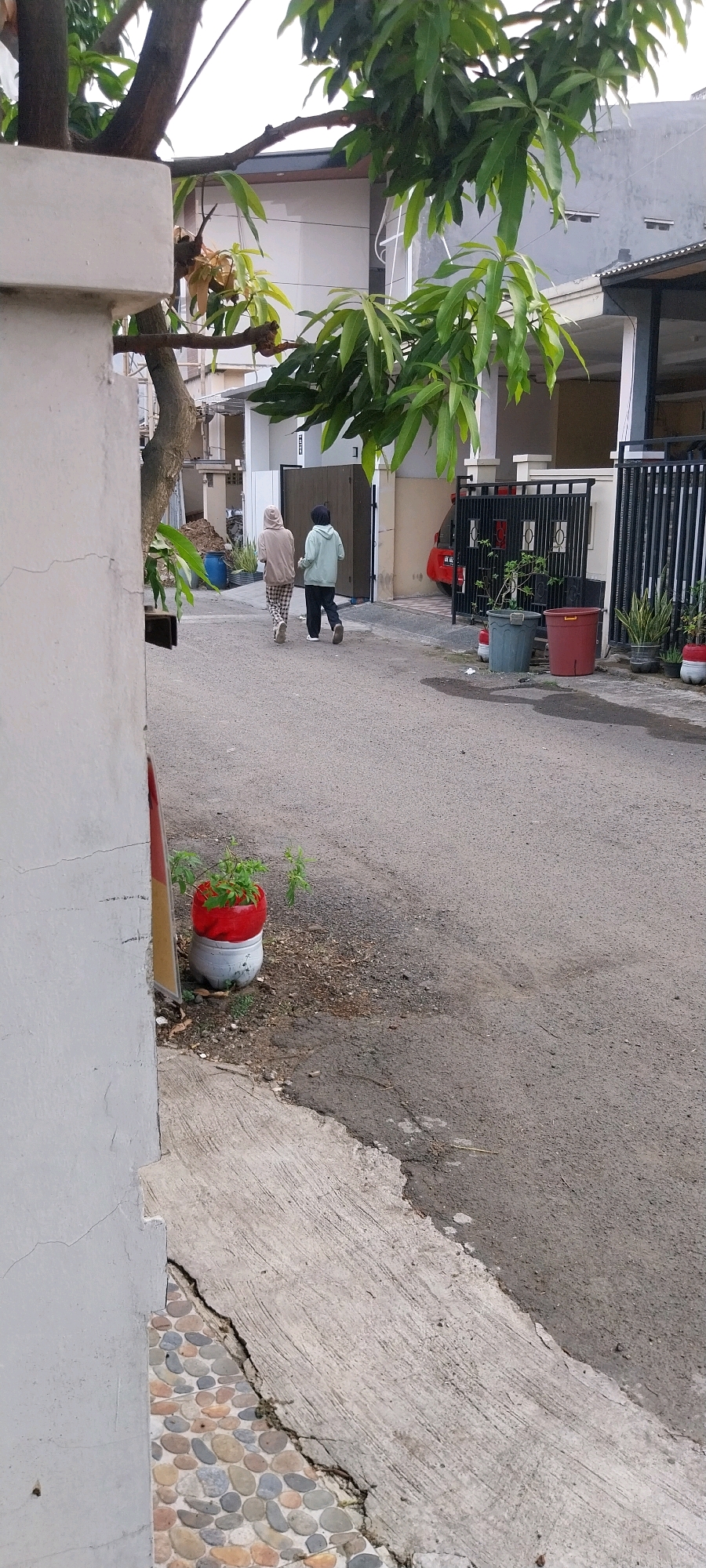 Two individuals walk down a paved street in Bojong Nangka, Indonesia. The person on the left wears a light brown hooded sweatshirt and plaid pants, while the person on the right is in a light green hooded sweatshirt and dark pants. They are walking away from the camera towards a closed gate with decorative vertical bars, a white house, and a red car visible behind it. To the left of the street, a white concrete wall and a tree with green leaves are prominent. In the foreground, on the left, a plant is in a white pot with a red top, and further down the street, there are two larger planters, one gray and one red. The environment appears to be a residential street with houses and fencing.