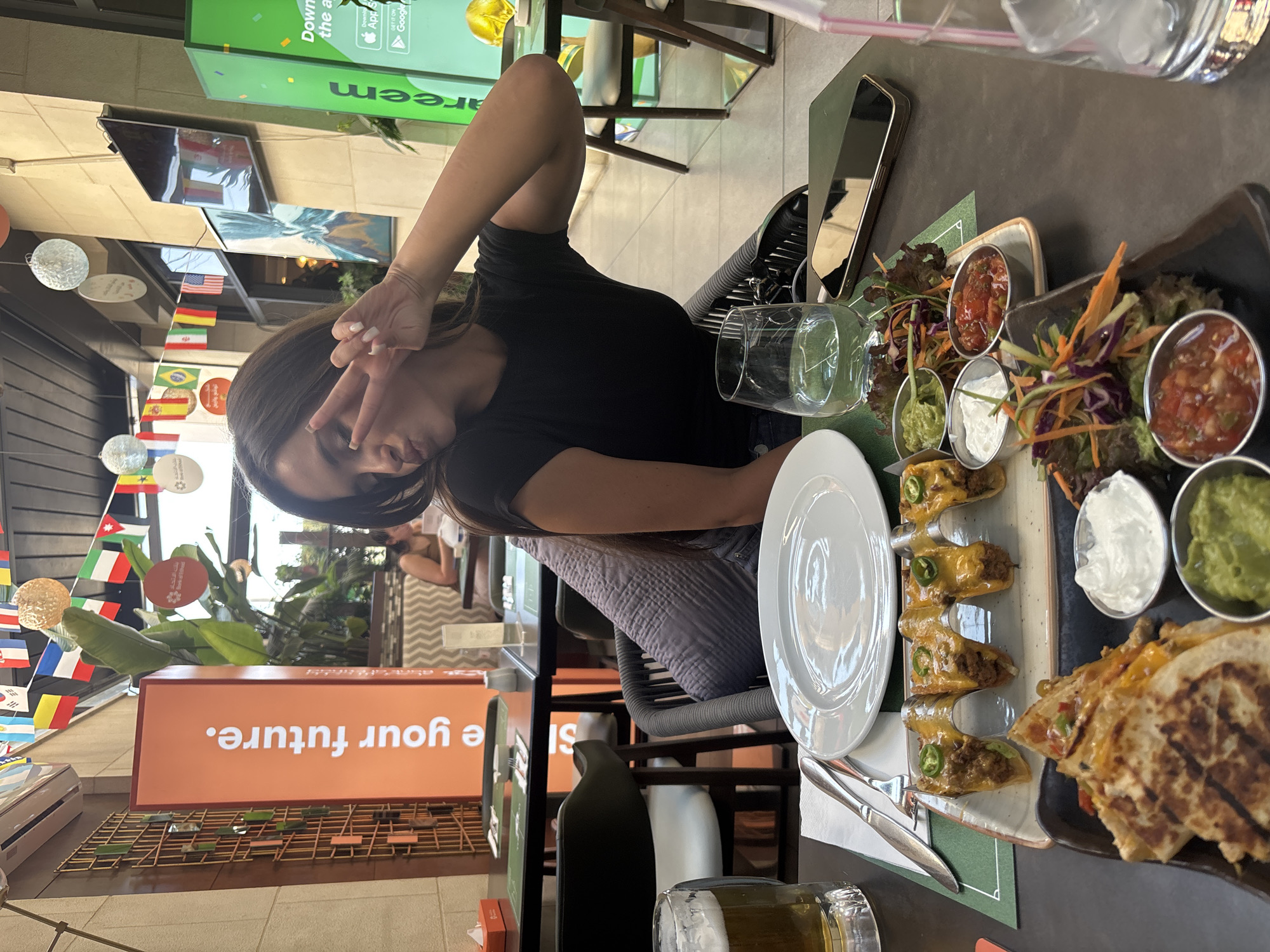 A young woman with dark hair is seated at a table in an outdoor dining area, making a peace sign and pouting at the camera. She is wearing a black t-shirt. In front of her is a table laden with food and drinks. A glass of beer is on the left, and a plate of quesadillas, tacos, guacamole, sour cream, and salsa is in the foreground. A glass of water sits on the table, along with a smartphone. Behind her, flags from various countries are strung across the ceiling, and banners with Arabic text are visible. The setting appears to be a restaurant in Amman, Jordan, indicated by the Arabic writing on a banner.