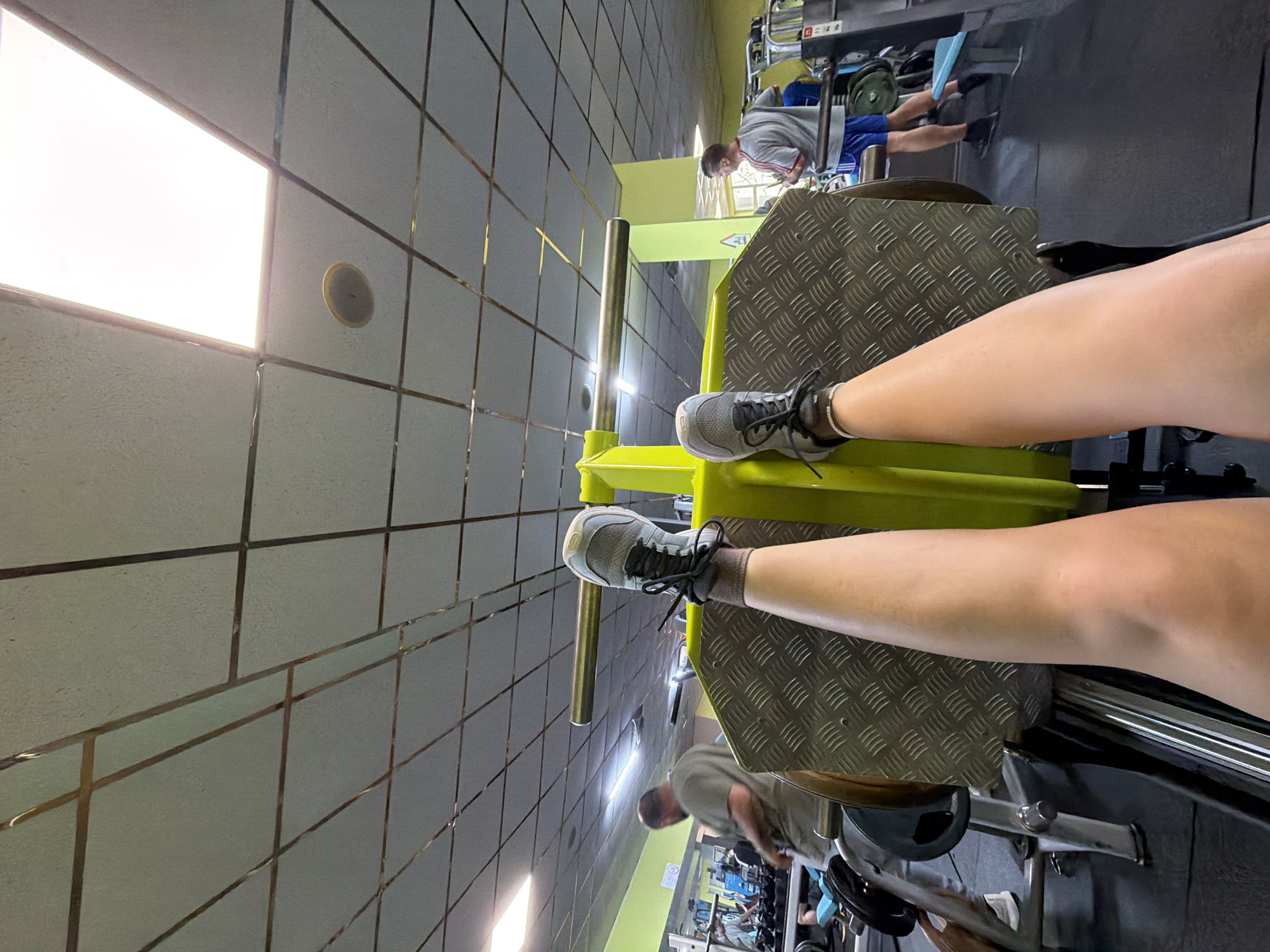 Here is a description of the image:
The image shows a person's legs extended on a leg press machine in a gym. The person is wearing grey sneakers. The gym has tiled walls and bright overhead lighting. Other gym equipment and people can be seen in the background. While the image was captured in a gym, the information to ascertain that it is located in Varna, Bulgaria is not provided. Therefore, it is impossible to provide that information.