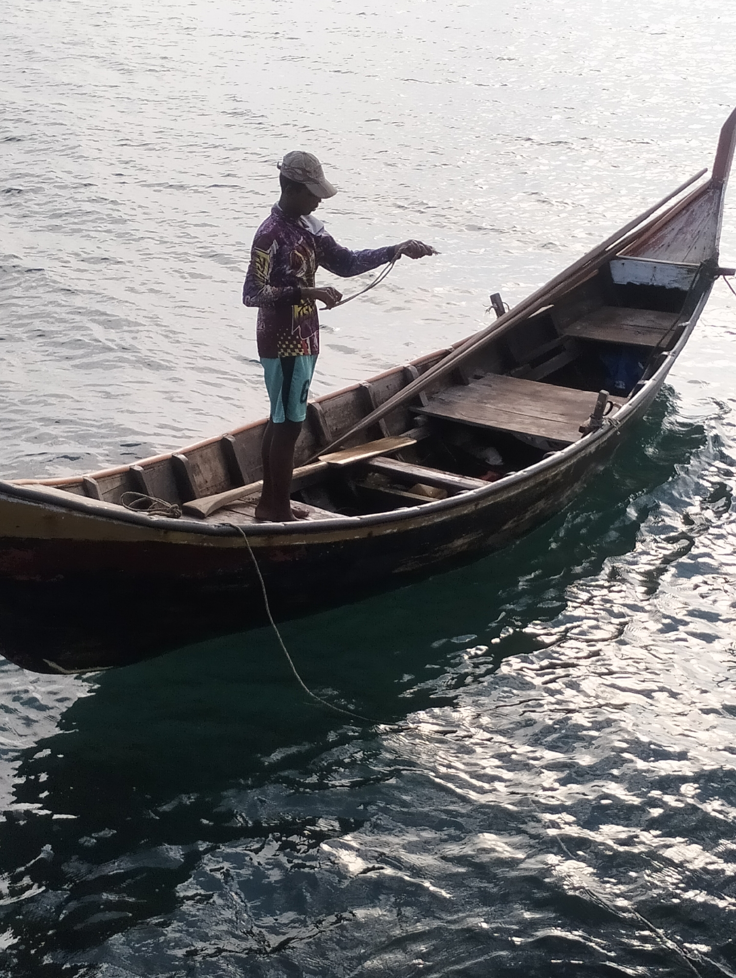 A man stands in a long, narrow, wooden boat on the water. He is wearing a cap, a long-sleeved patterned shirt, and shorts. He appears to be holding a rope. The boat is dark-colored and has wooden planks inside. The water is rippled and reflects the light. The location is Lhokseumawe, Indonesia.