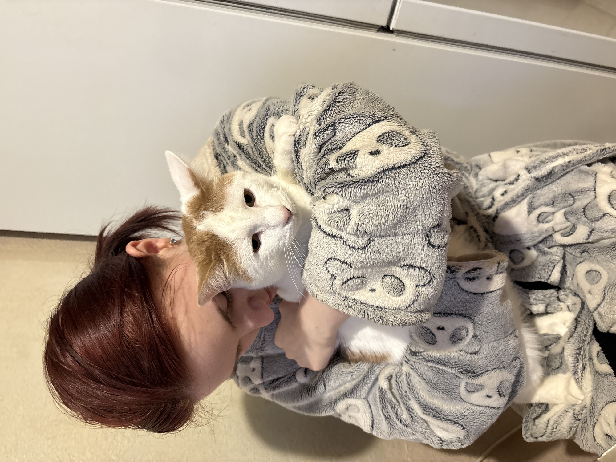 A young woman with auburn hair pulled back in a ponytail is holding a white and orange cat. She is wearing a grey fleece robe with a pattern of pandas. The woman is looking down at the cat, and her face is partially obscured by the cat's head. The cat appears calm and is nestled in her arms. The background is a plain wall and a white vertical surface, possibly a door or cabinet. The scene suggests a domestic setting.